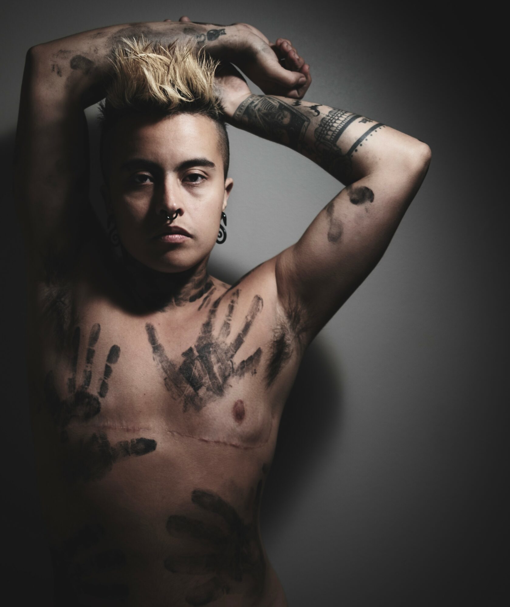 Person with tattoos and short blonde hair poses with arms above head, looking at camera, dark background.