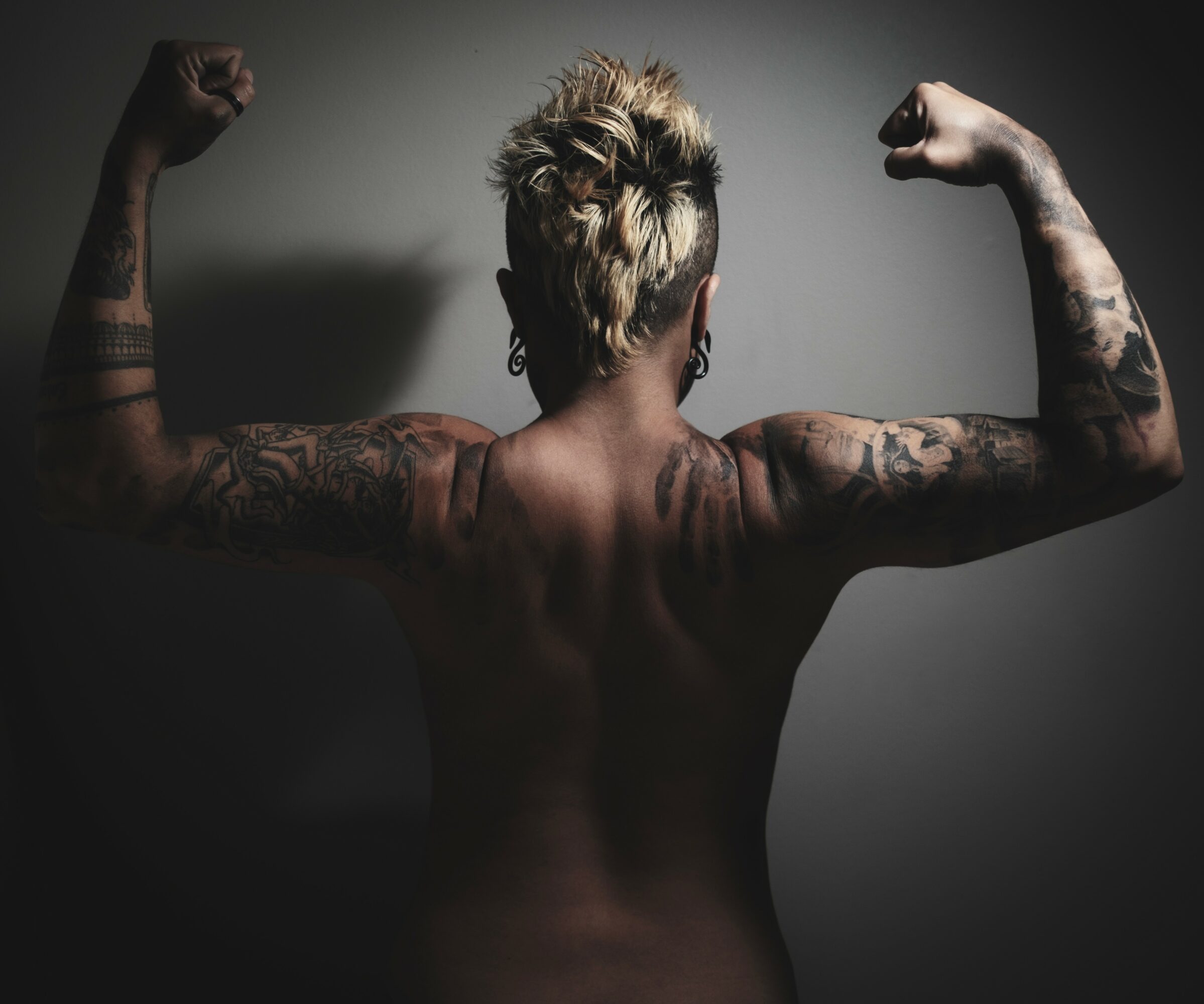 Back view of a person with short, spiky hair, flexing arms to show tattoos on shoulders and upper arms.