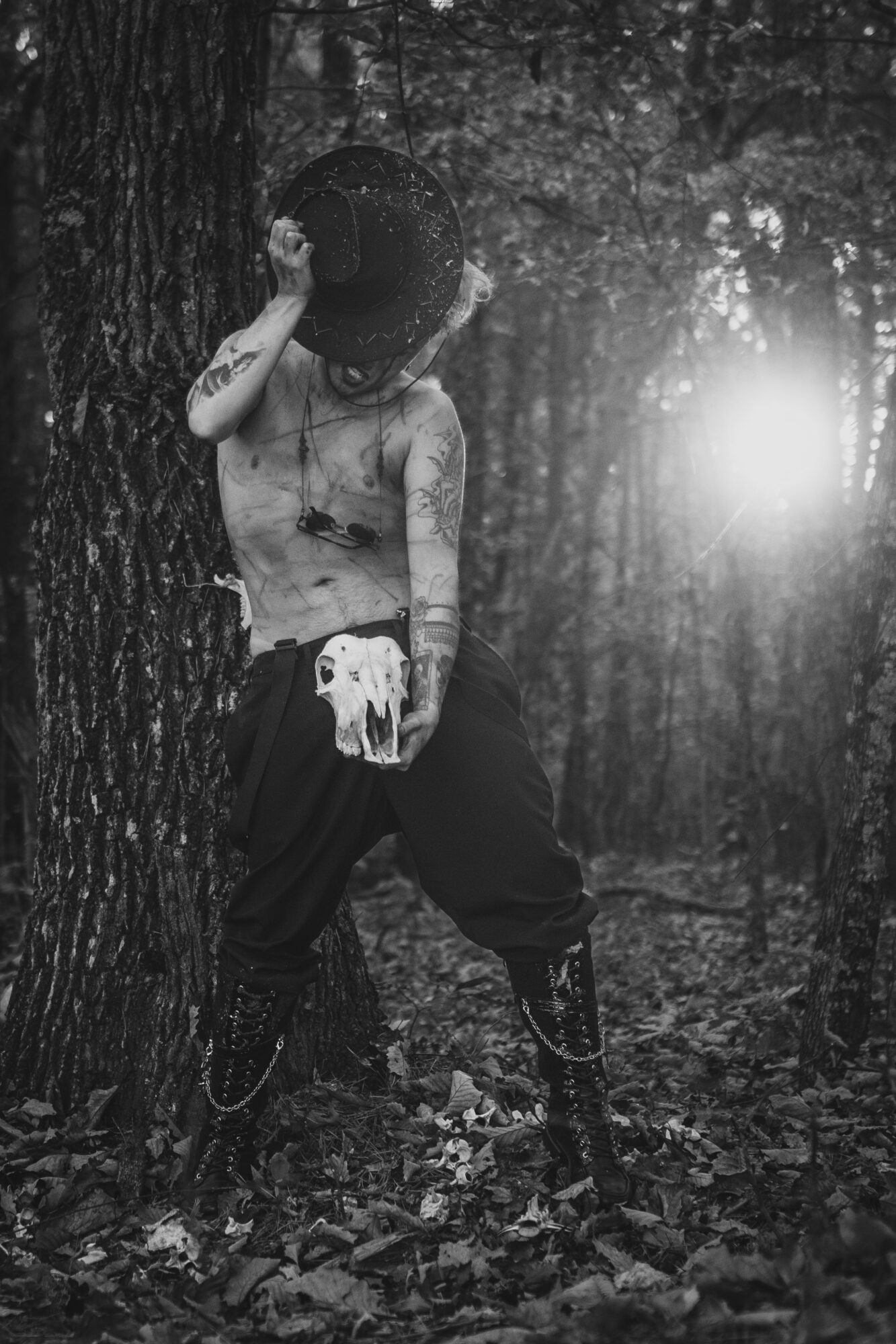 Child standing in a forest holding a hat, with a bright light in the background, wearing dark pants and no shirt.
