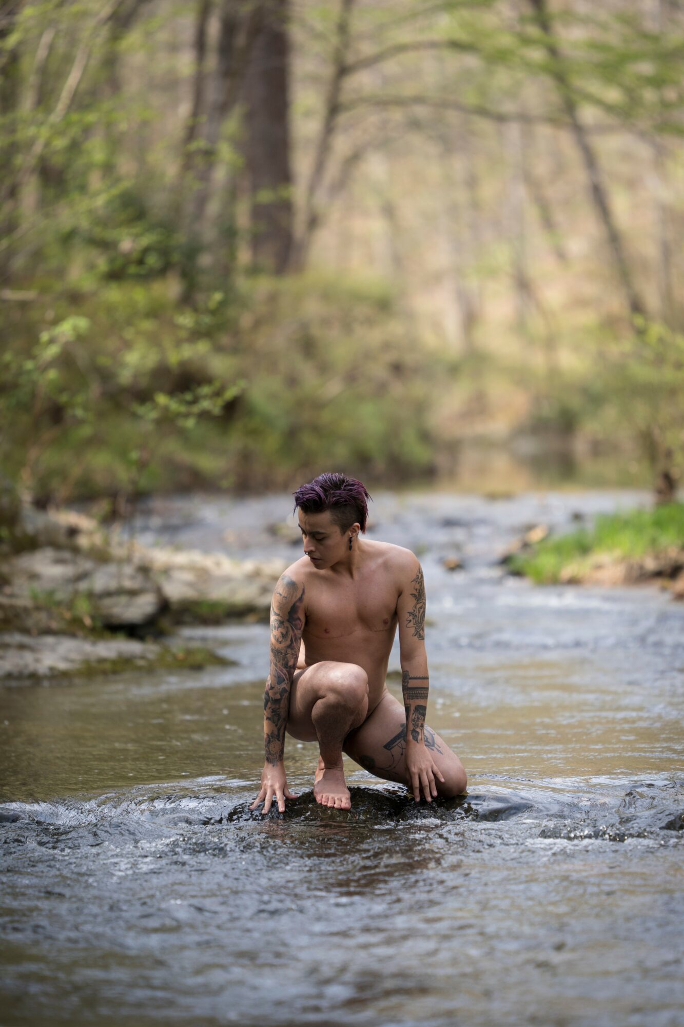 Person crouching in a shallow river surrounded by trees and greenery, with a natural outdoor setting.