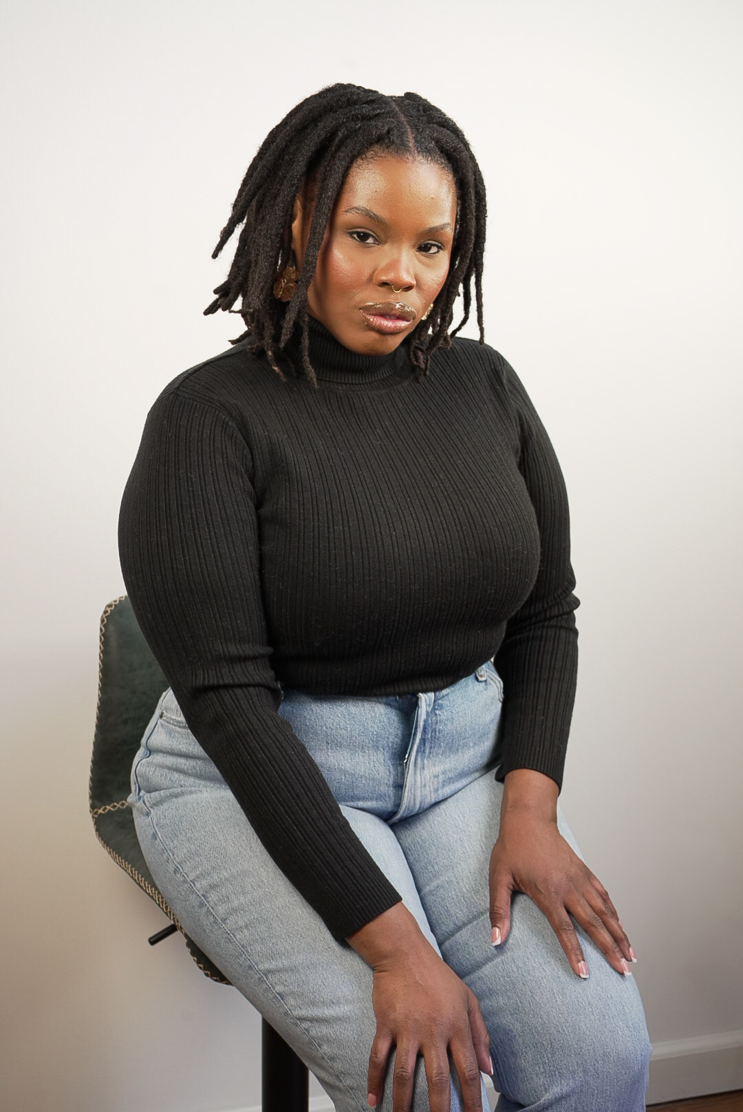 Woman with dreadlocks wearing a black sweater and light jeans, sitting on a chair against a plain background.