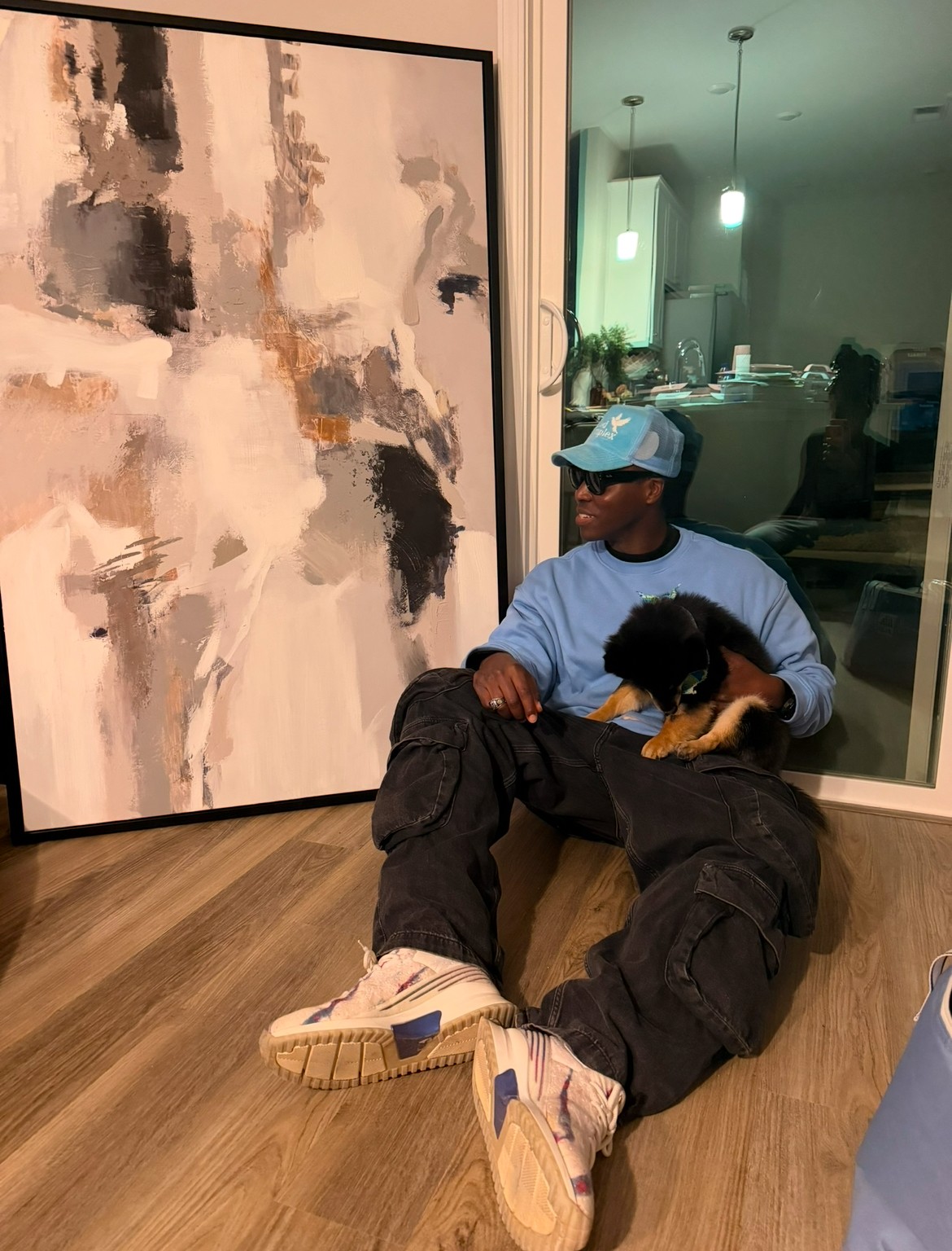 Person sitting on the floor holding a small dog, with a large abstract painting behind them and a dining area visible through a window.