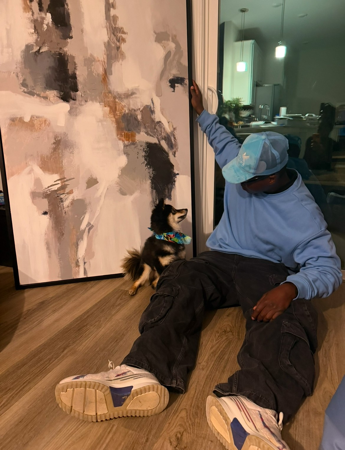 Person sitting on the floor with a dog, reaching towards a large abstract painting, in a room with a window and kitchen.