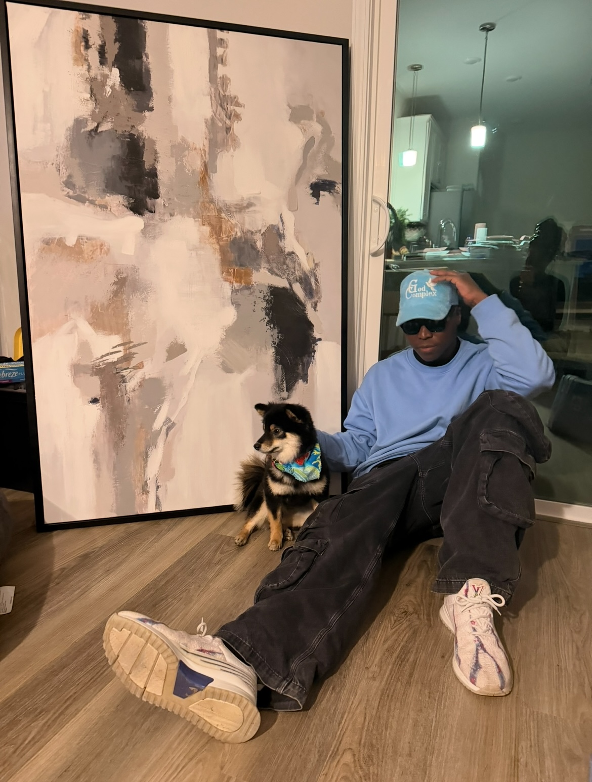 Person sitting on the floor with a dog, large abstract painting behind them, in a room with wooden floor and modern lighting.