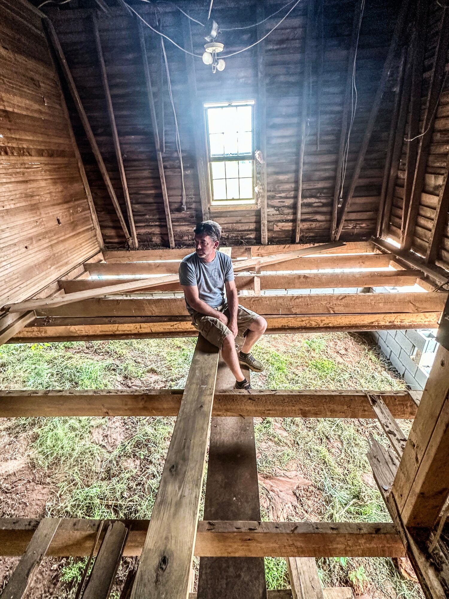 Person sitting on a wooden beam inside an unfinished attic with a window, wooden walls, and exposed beams.