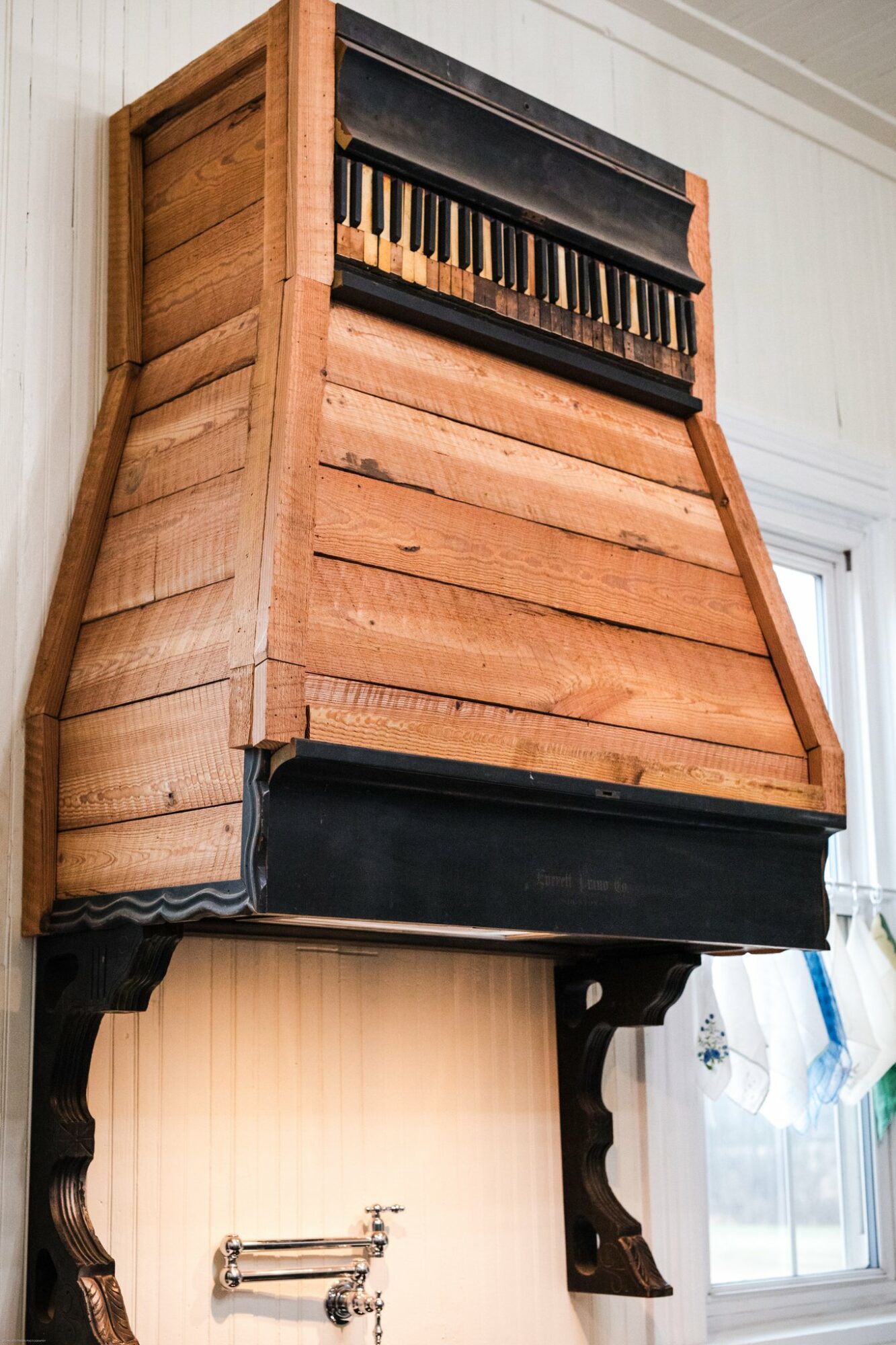 Upright wooden cabinet with a small keyboard on top, black base, and decorative legs, next to a window.