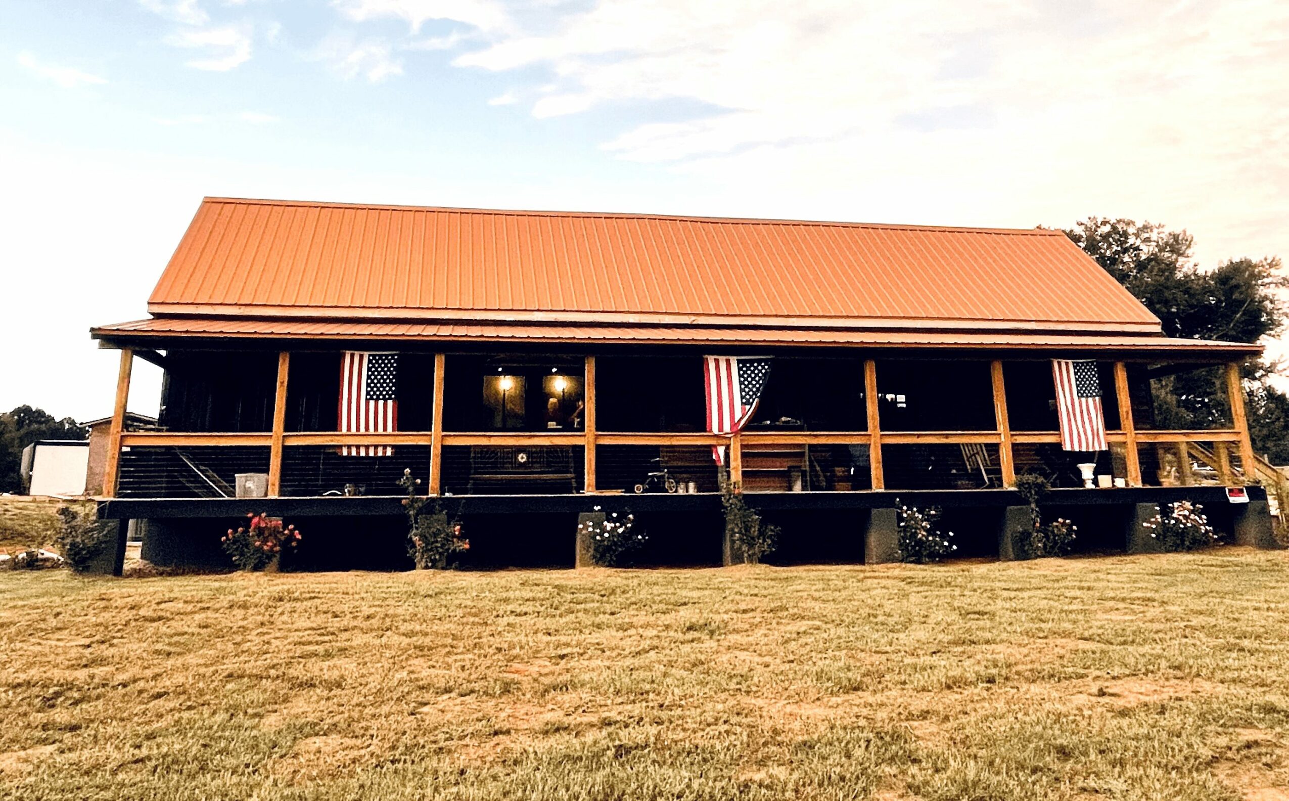 Long building with a red roof, wooden porch, and American flags, set on a grassy area with trees in the background.
