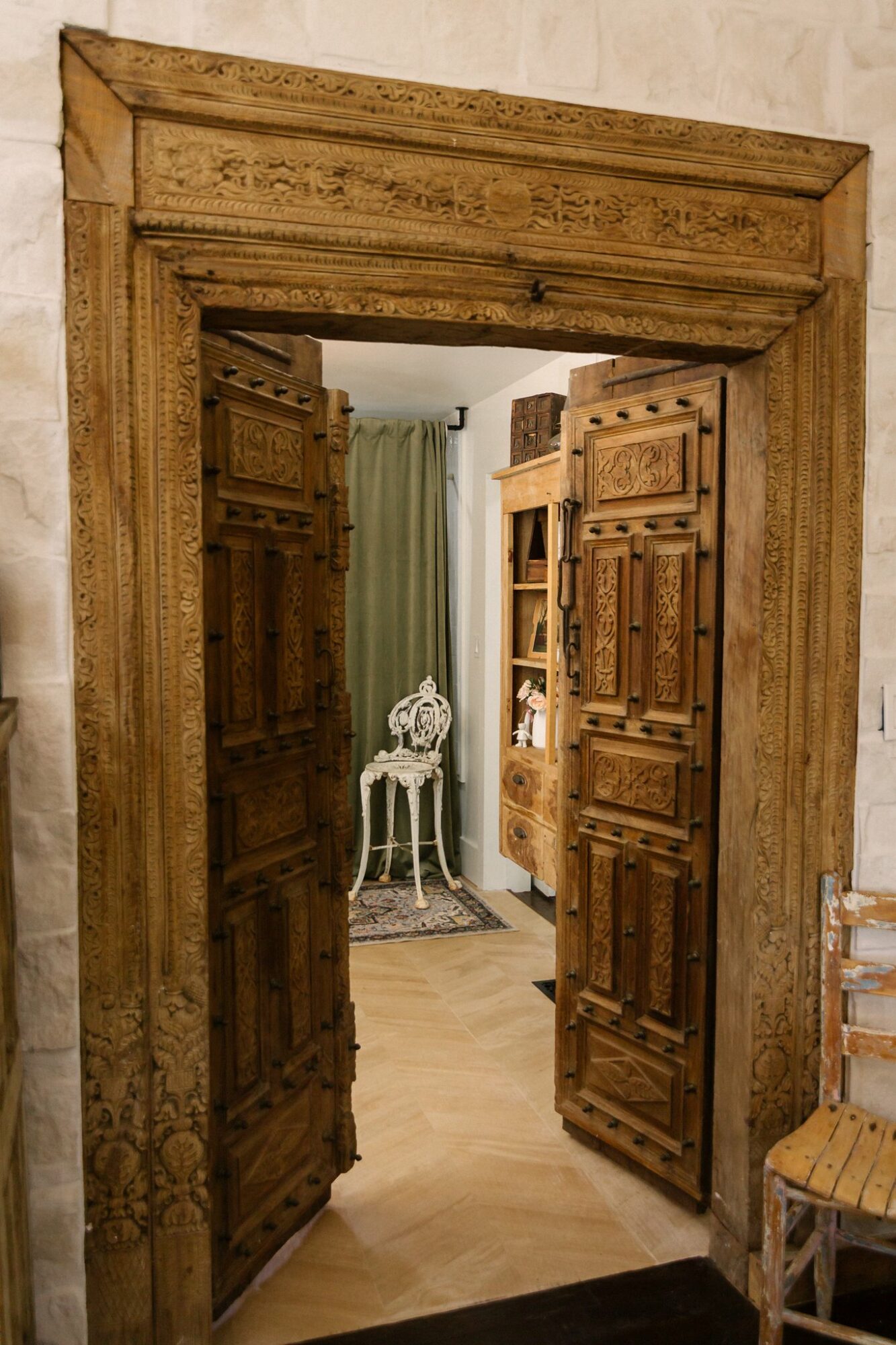 View through an open wooden door into a room with a small table and a rug, with curtains and bookshelves visible.