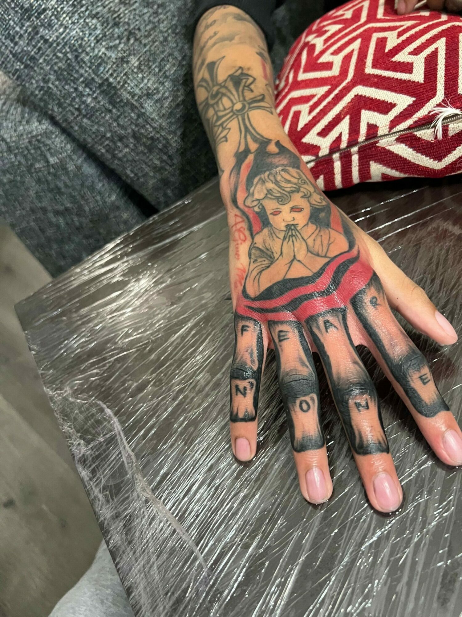 Tattooed hand with fingers spread, resting on a plastic-covered surface, wearing a red and white patterned sleeve.
