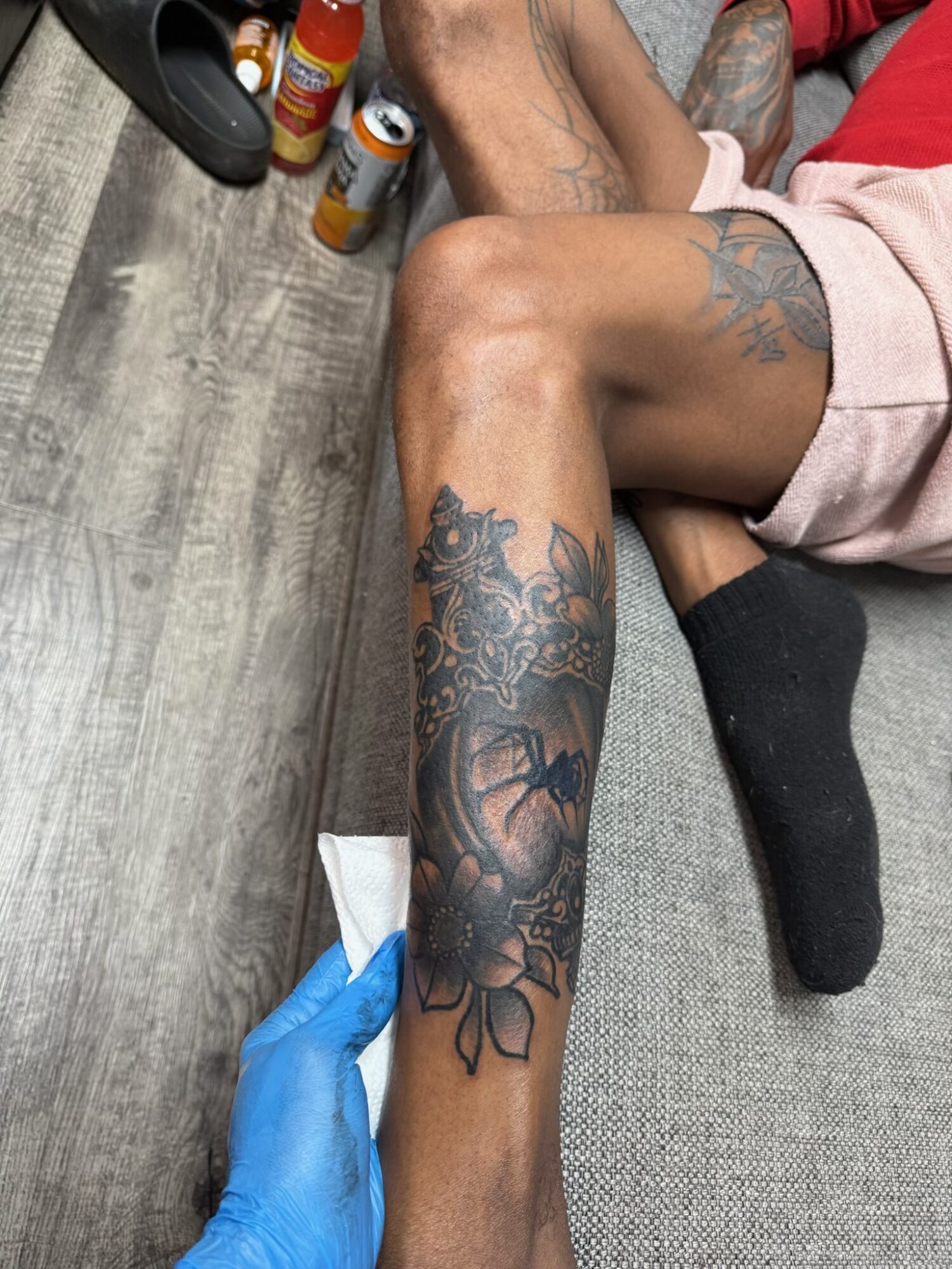 Person lying on side with tattoos on leg, wearing black sock, with a gloved hand holding a tool near the leg, on a wooden floor.