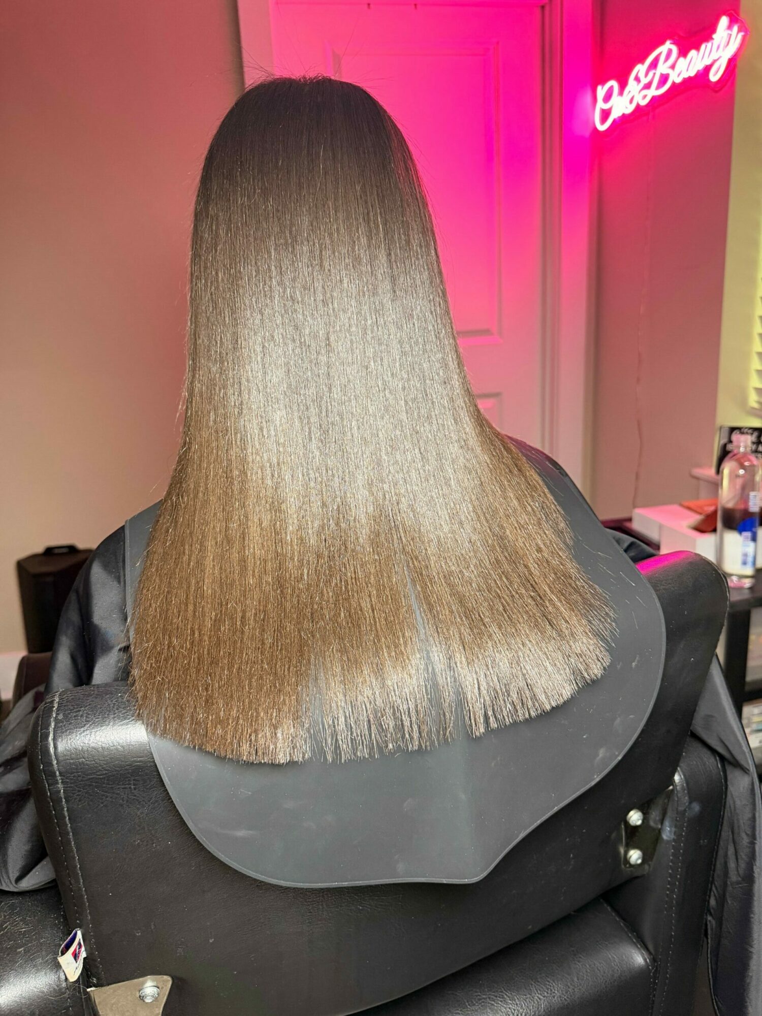 Back of a person with long, straight, light brown hair sitting in a salon chair, pink neon light in background.