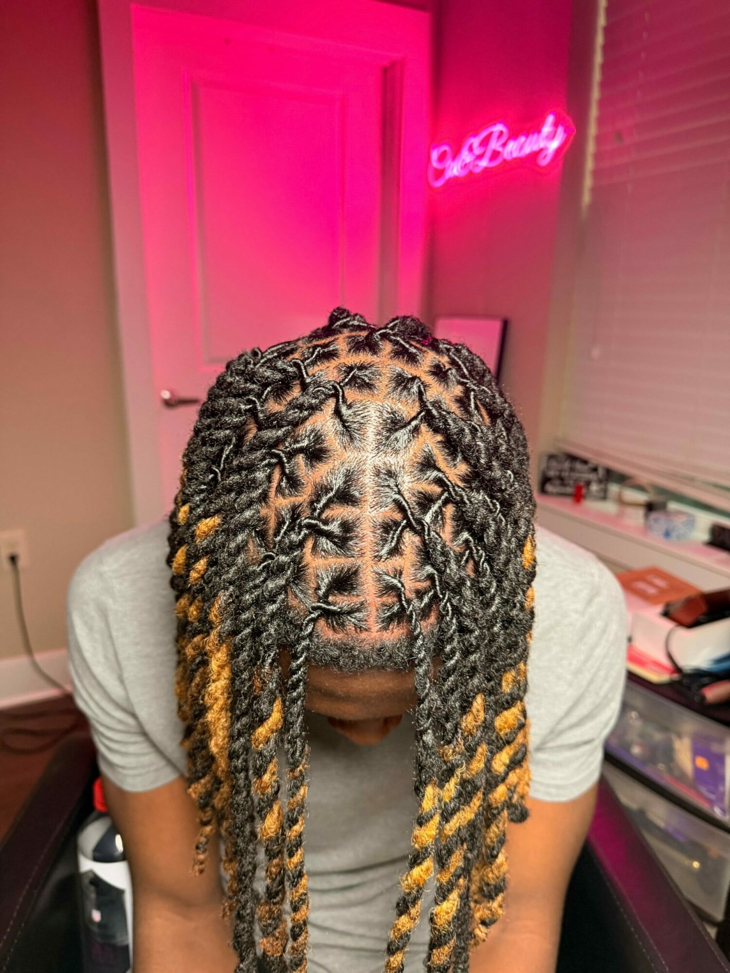 Person with braided hair in a room with pink lighting and a neon sign, wearing a gray shirt.