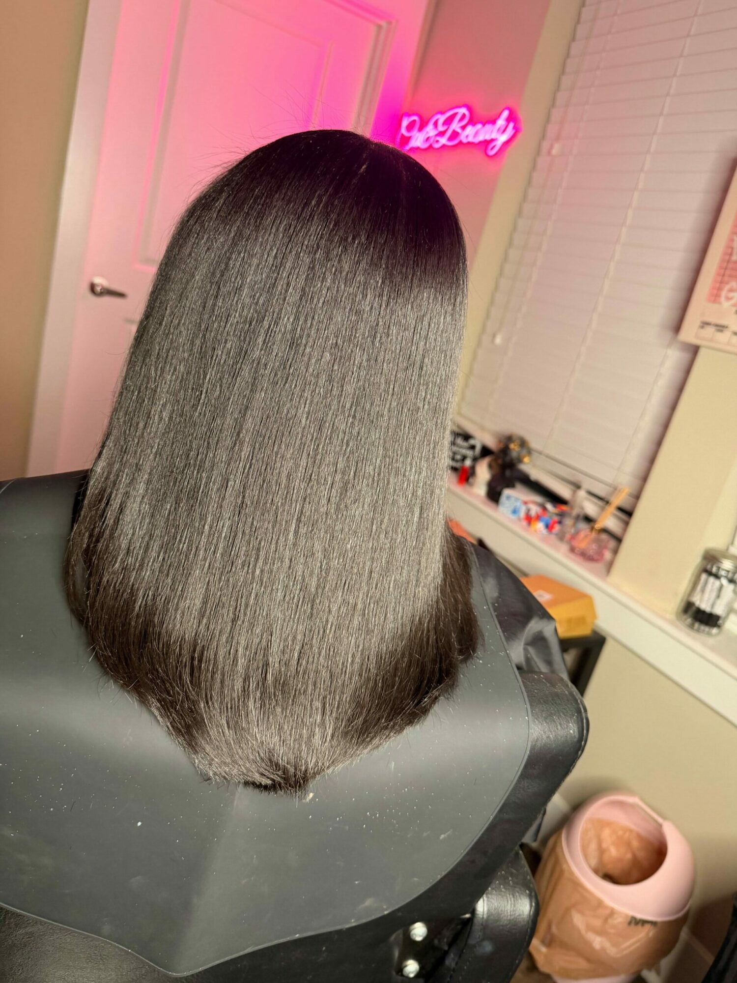 Person with straight, shiny, shoulder-length hair sitting in a salon chair, with salon equipment and pink neon sign in background.
