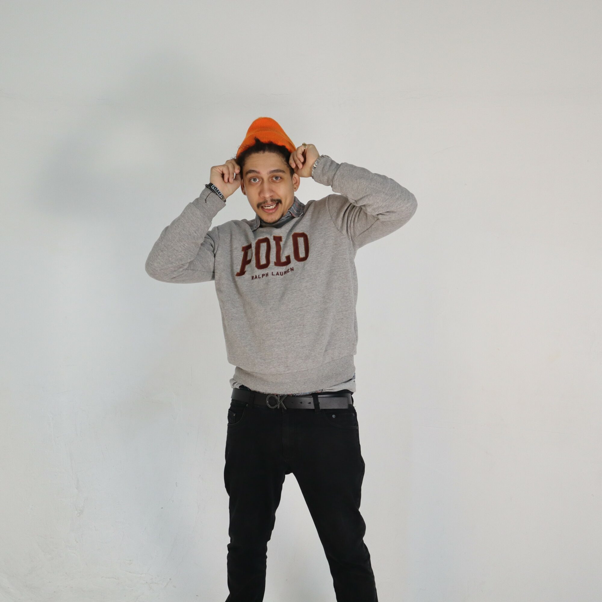 Person wearing a gray POLO sweatshirt and orange hat, holding the hat with both hands, smiling, standing against a plain background.