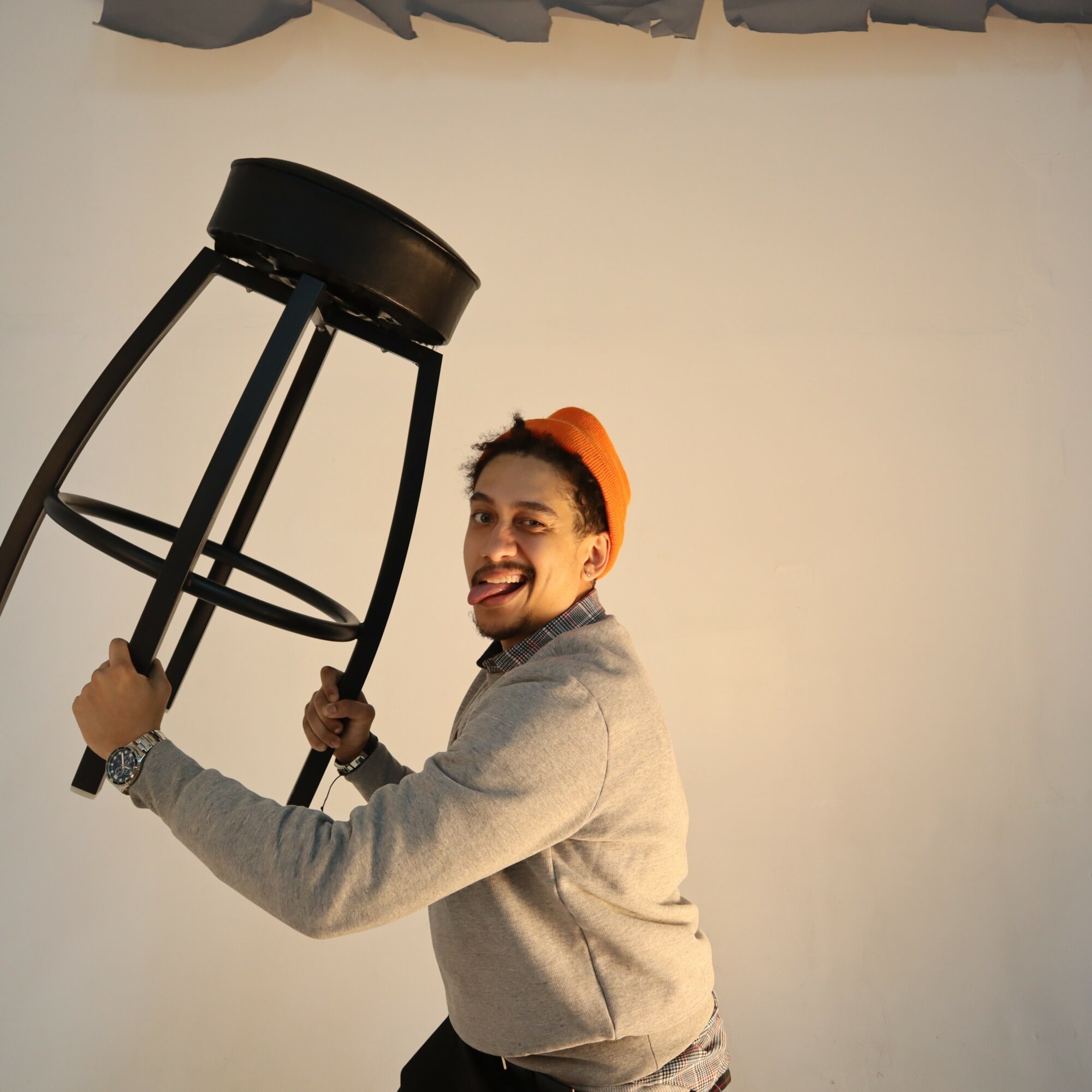 Person holding a stool, smiling, wearing a gray hoodie and orange cap, against a plain background.