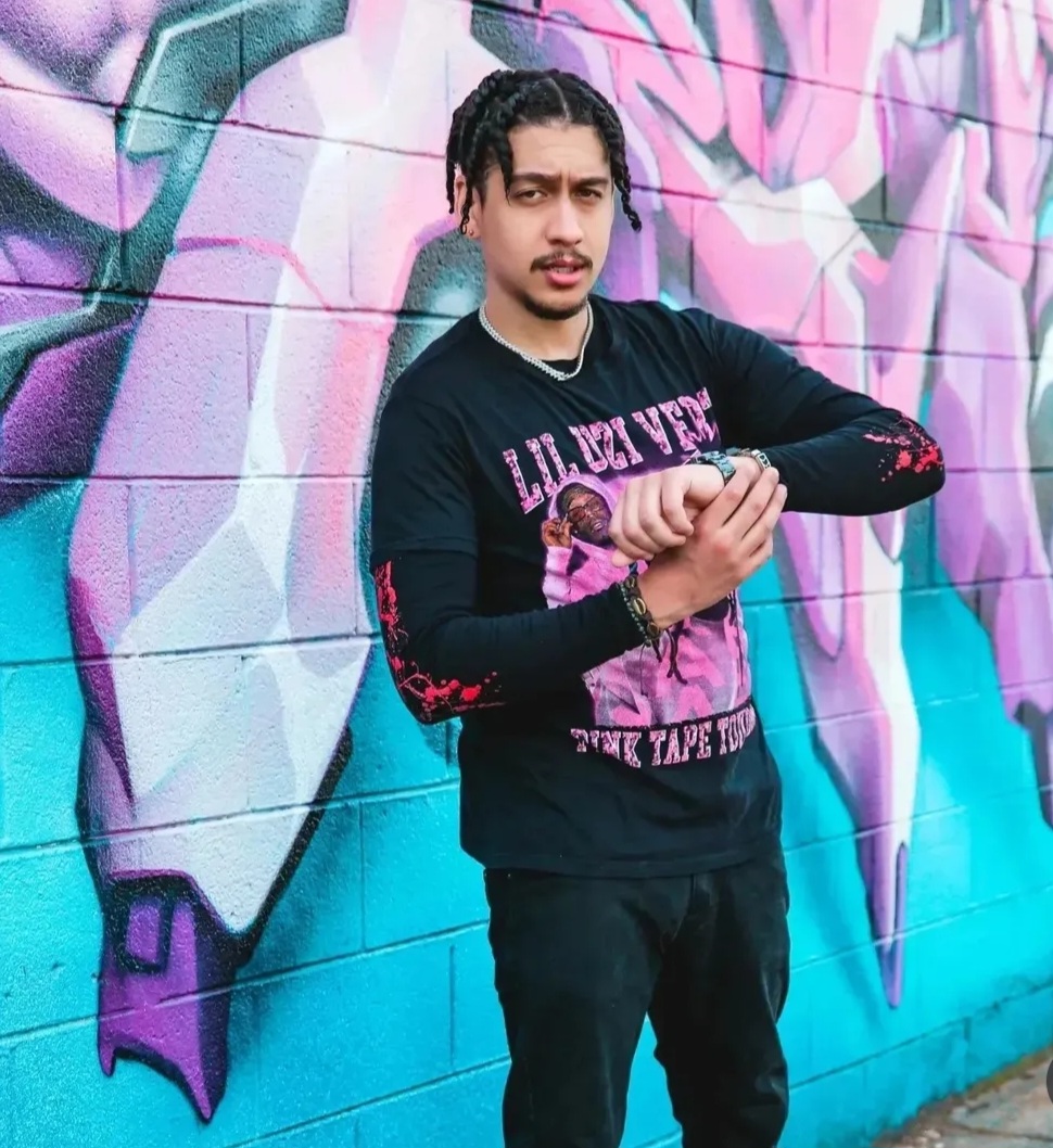 Young man with braided hair stands in front of colorful graffiti wall, wearing a black graphic sweatshirt and black pants.