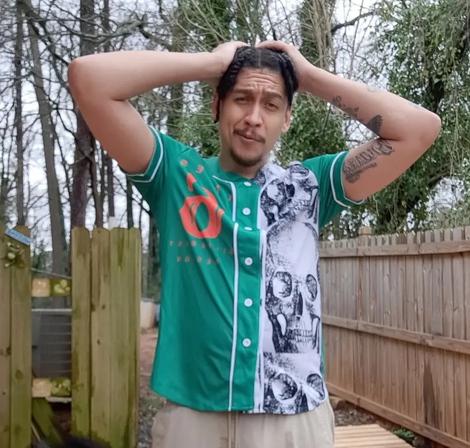 Young man with tattoos, wearing a green and white shirt, standing outdoors with trees and a wooden fence behind him.