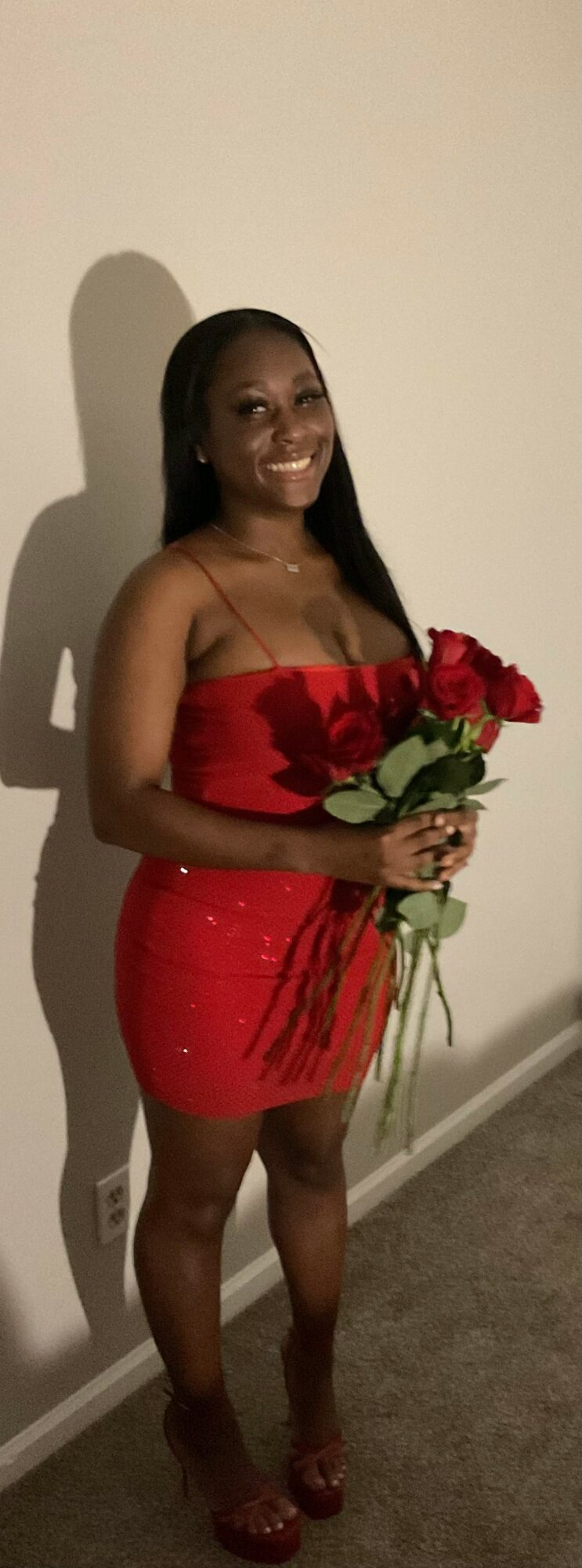 Woman in red dress holding a bouquet of red roses, smiling, standing against a plain wall.