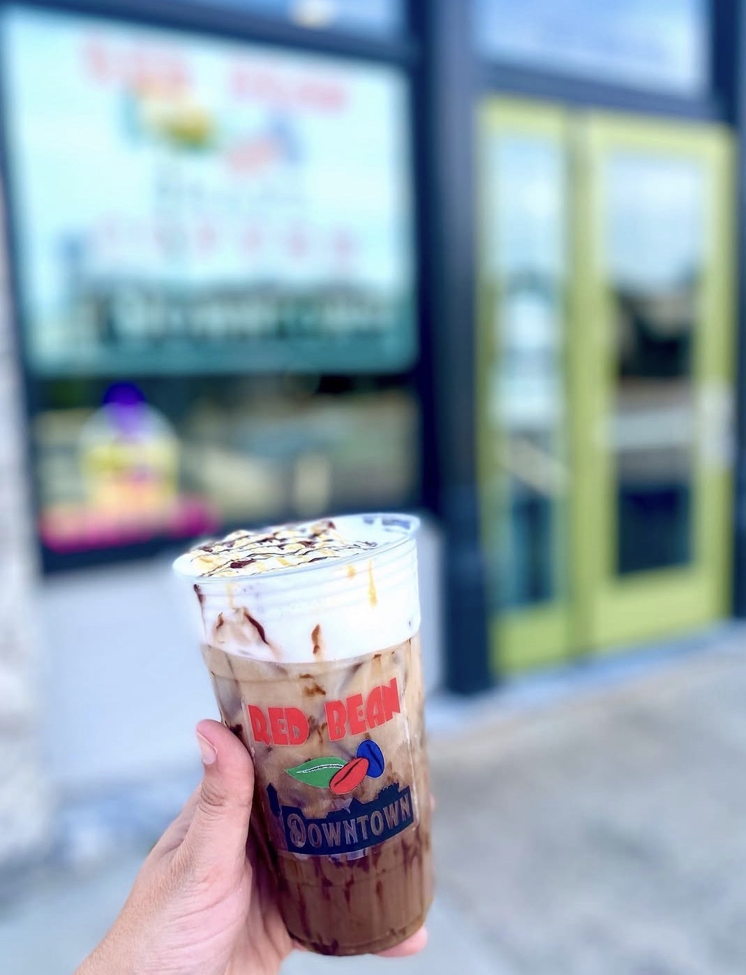 Hand holding a chocolate and vanilla layered drink with whipped cream outside a building with glass doors.