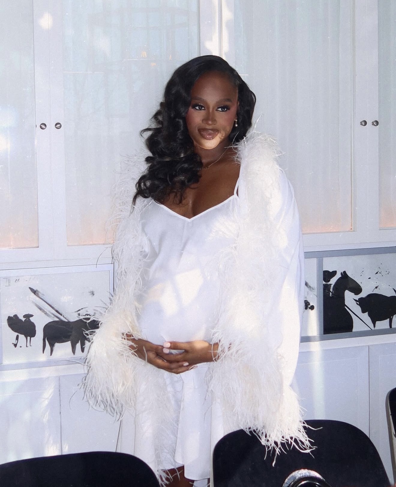 Woman in white dress and feathered shawl standing indoors with white cabinets and animal silhouettes in background.
