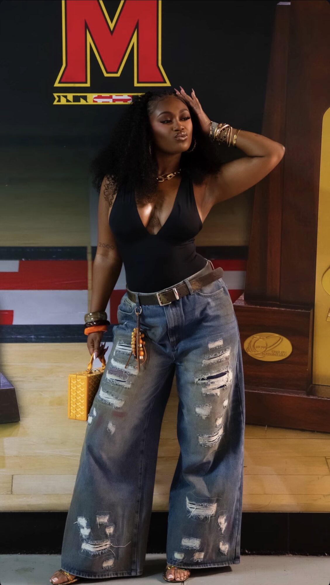 Woman with long curly hair wearing a black top and wide jeans, standing indoors with a colorful background.