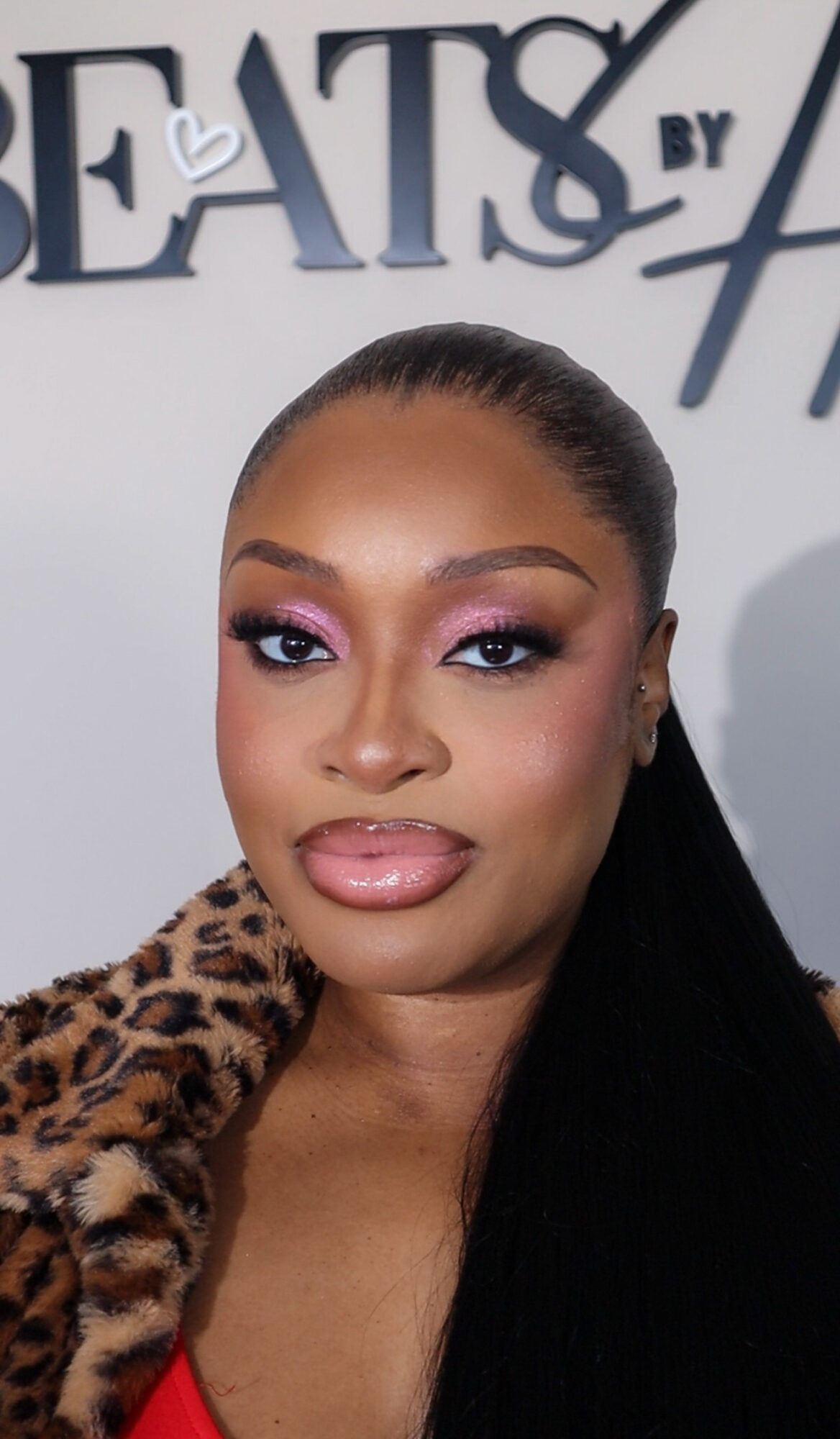 Close-up of a woman with makeup, wearing a leopard print garment, standing in front of a sign that says 'BEATS BY'.