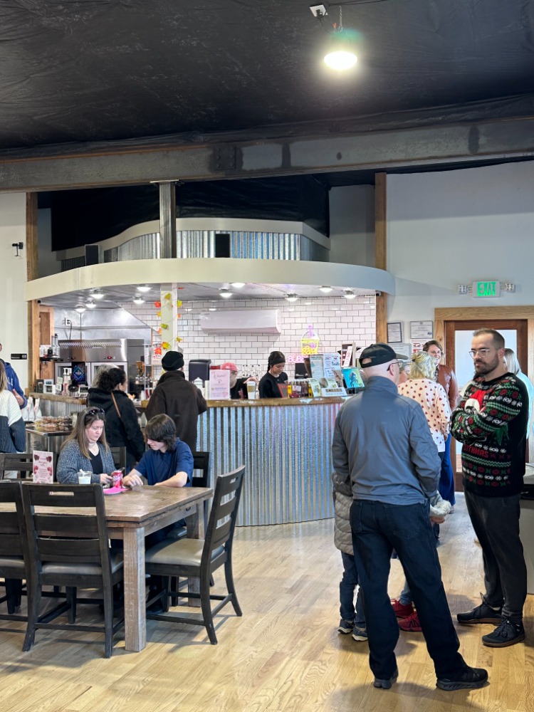 People gather in a cafe or restaurant with a counter and seating area, some standing and some sitting at tables.