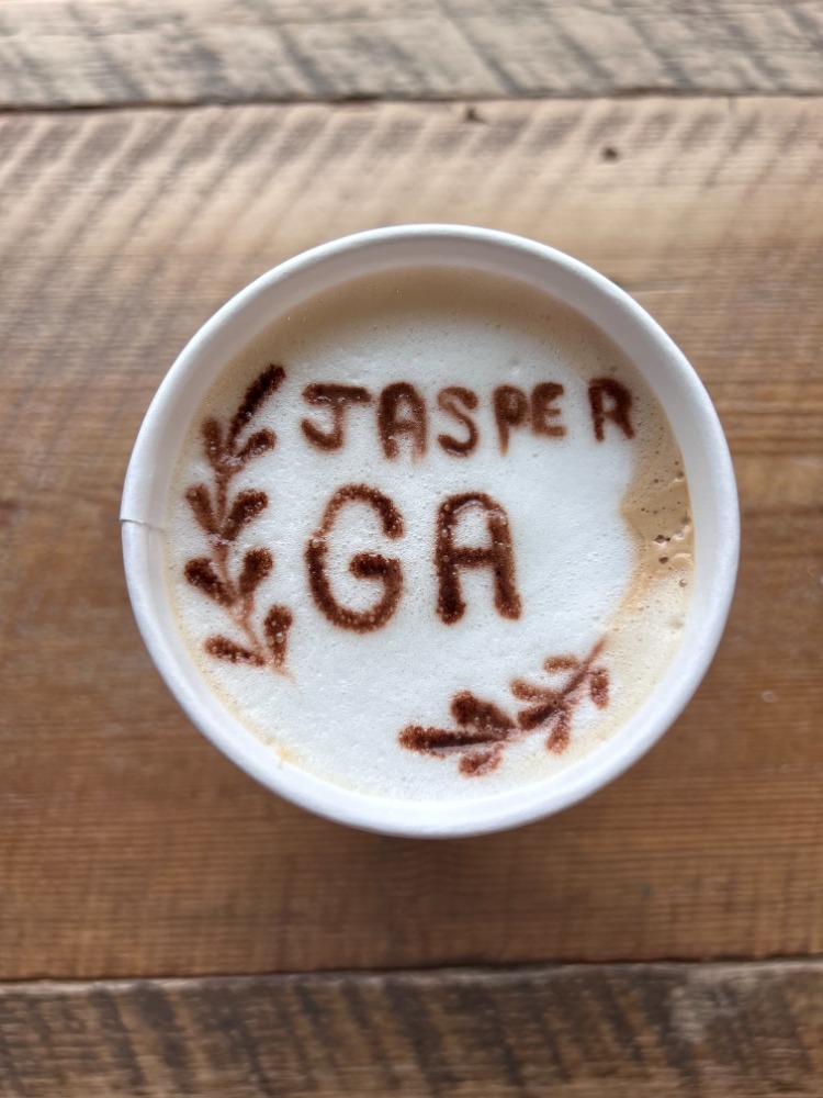 Top-down view of a cup of coffee with 'JASPER GA' written in cocoa powder and decorative leaves, on a wooden surface.