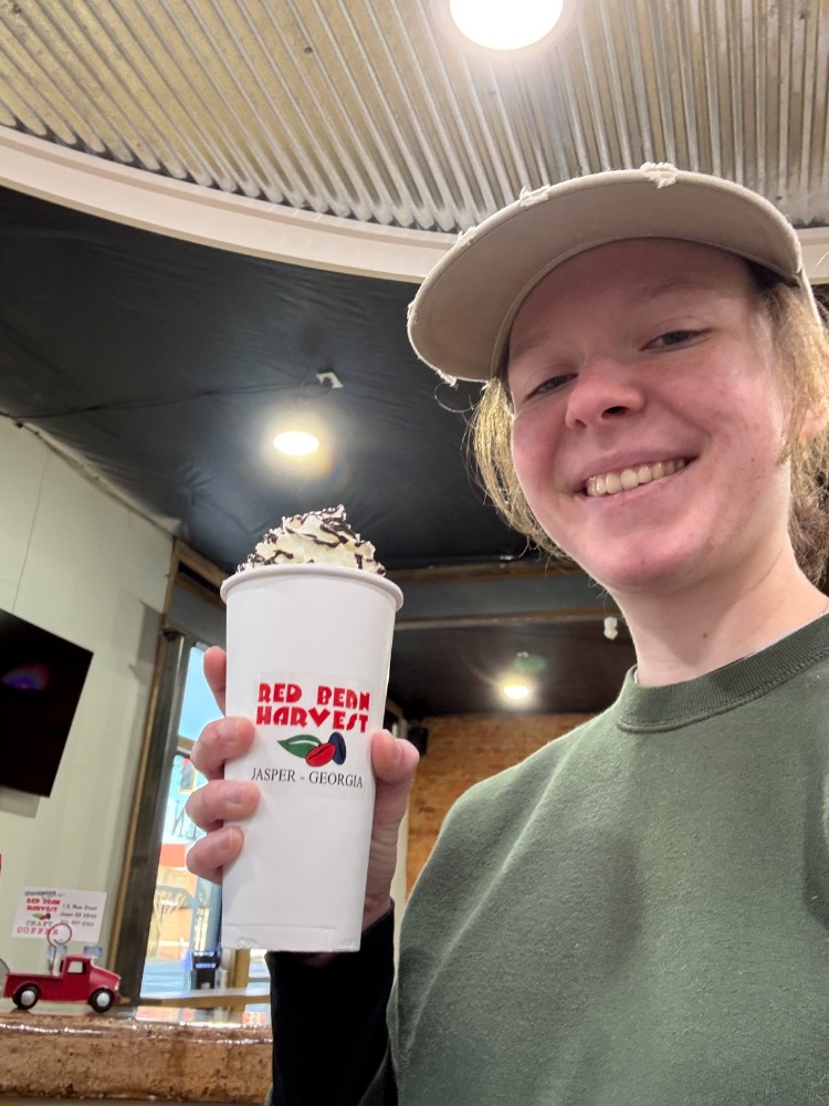 Person smiling and holding a large milkshake with whipped cream inside a fast-food restaurant, wearing a cap and green shirt.