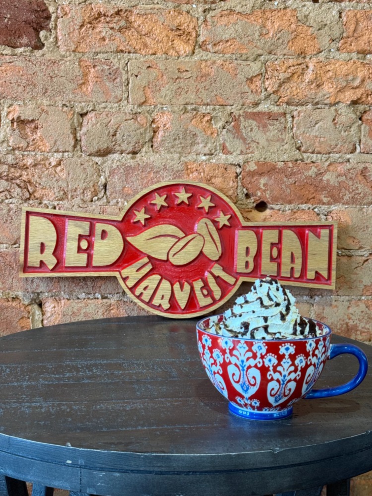 Red and white sign reading 'Red Bean' with a hand holding a bean, placed behind a blue cup filled with whipped cream on a wooden table.