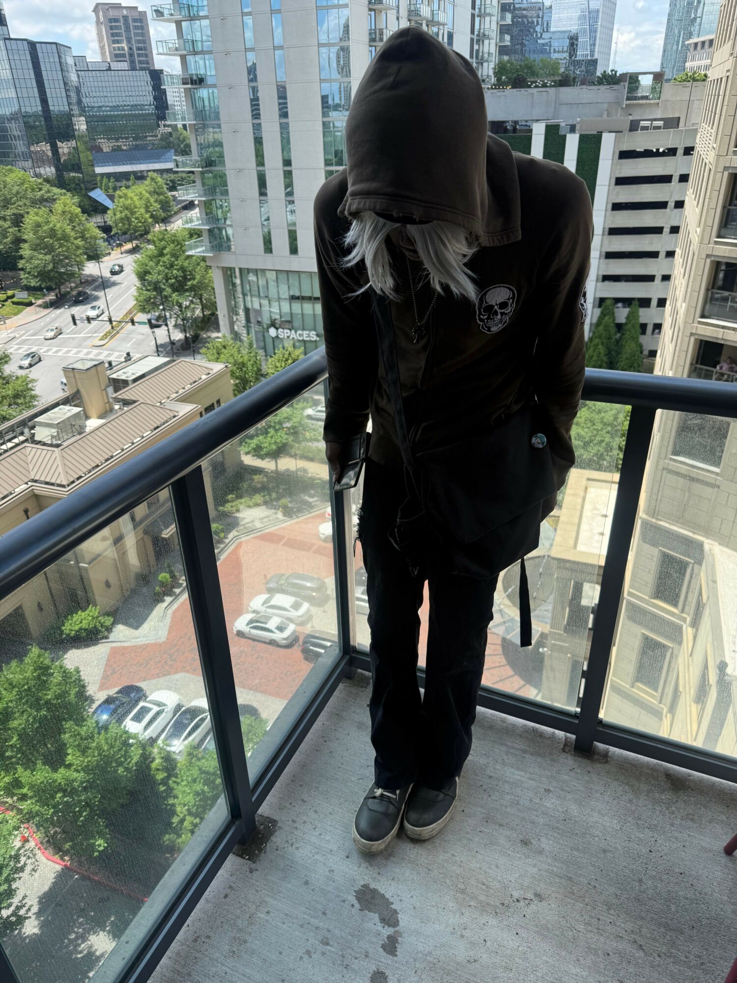 Person with long hair wearing dark clothing standing on balcony with city view behind glass railing.