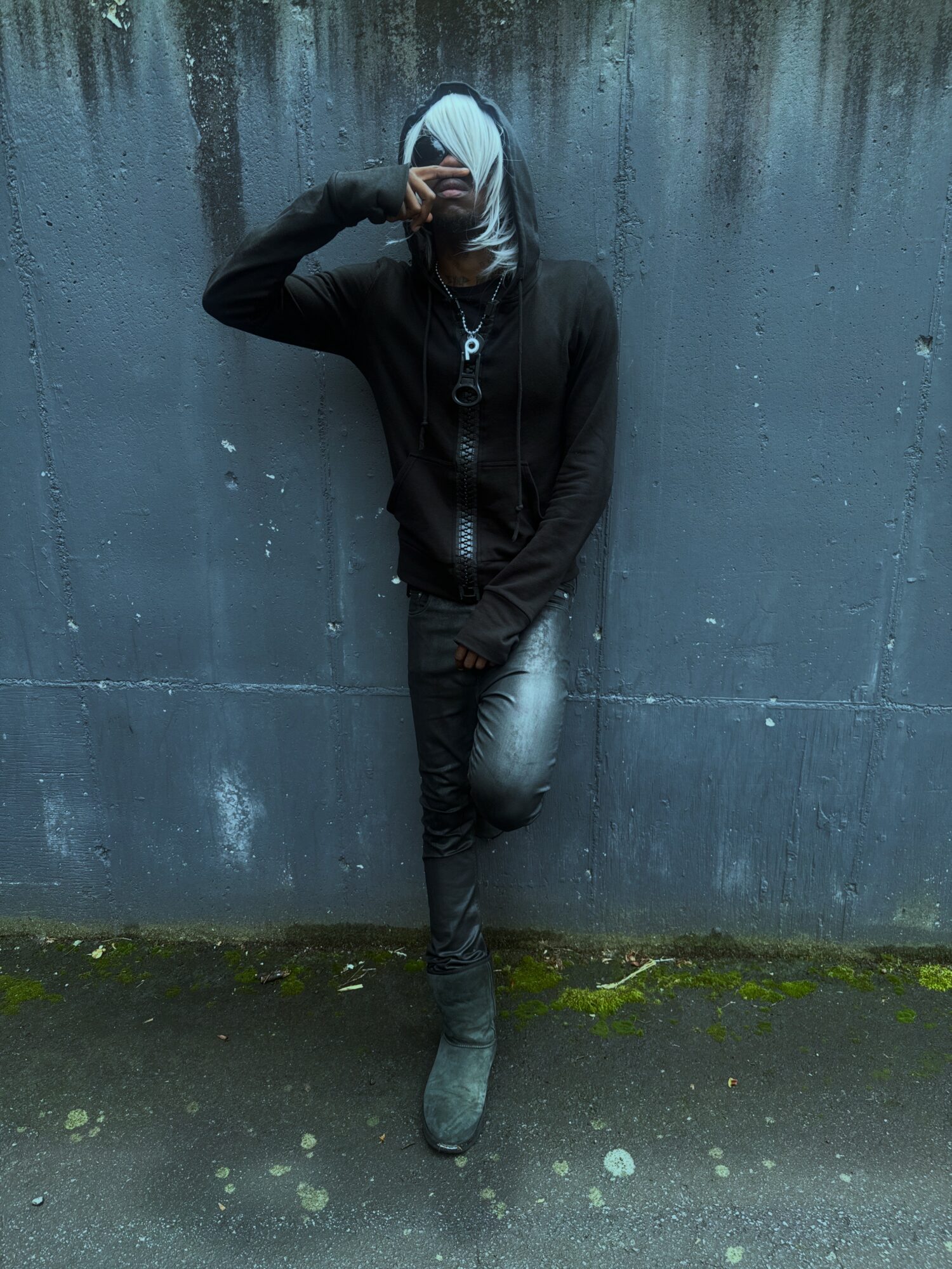 Person standing against a wall, wearing a hoodie, jeans, and boots, with one hand on their head, face covered with a scarf.