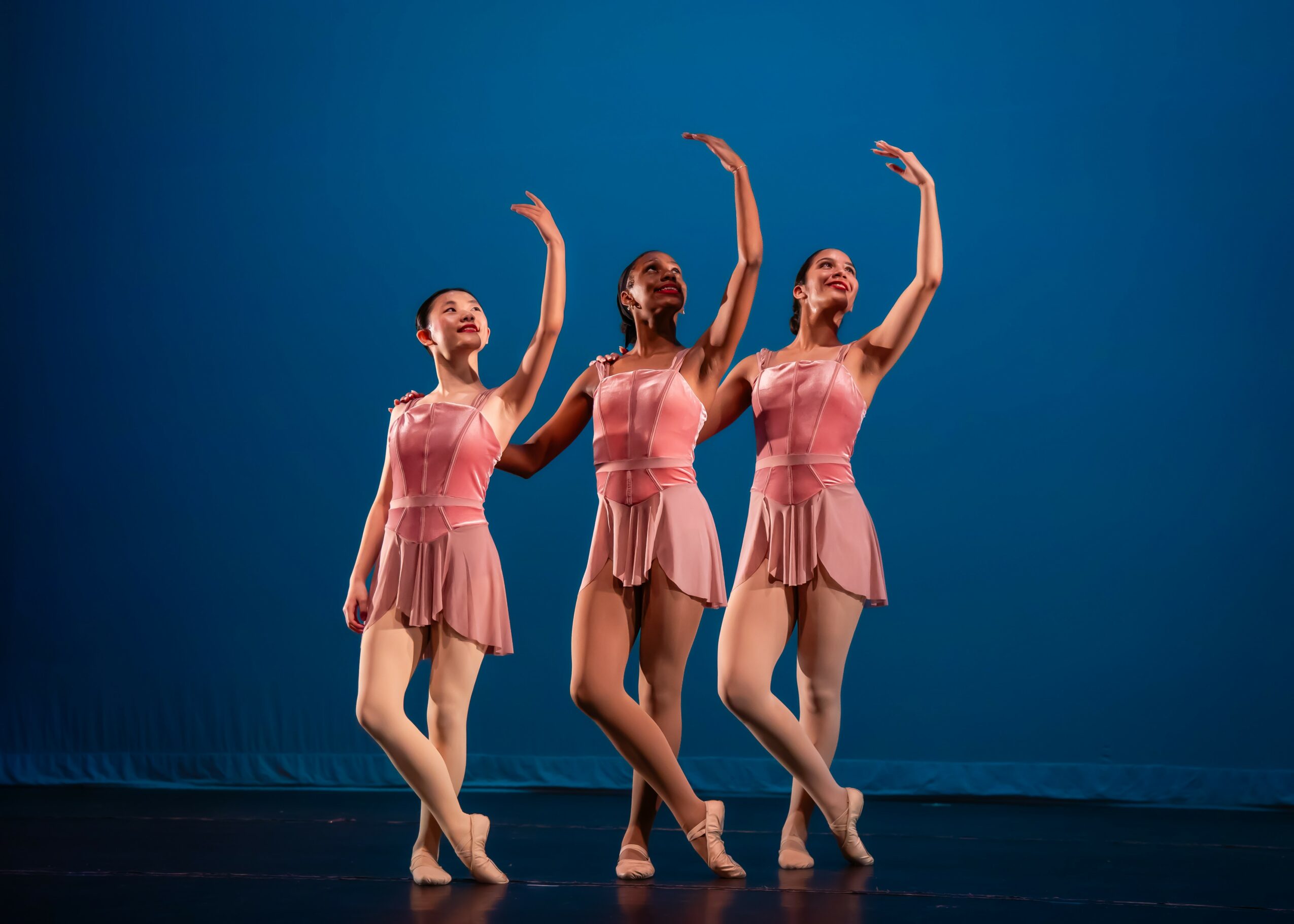 Three ballet dancers in pink dresses perform on stage with a blue background, arms raised gracefully, facing forward.