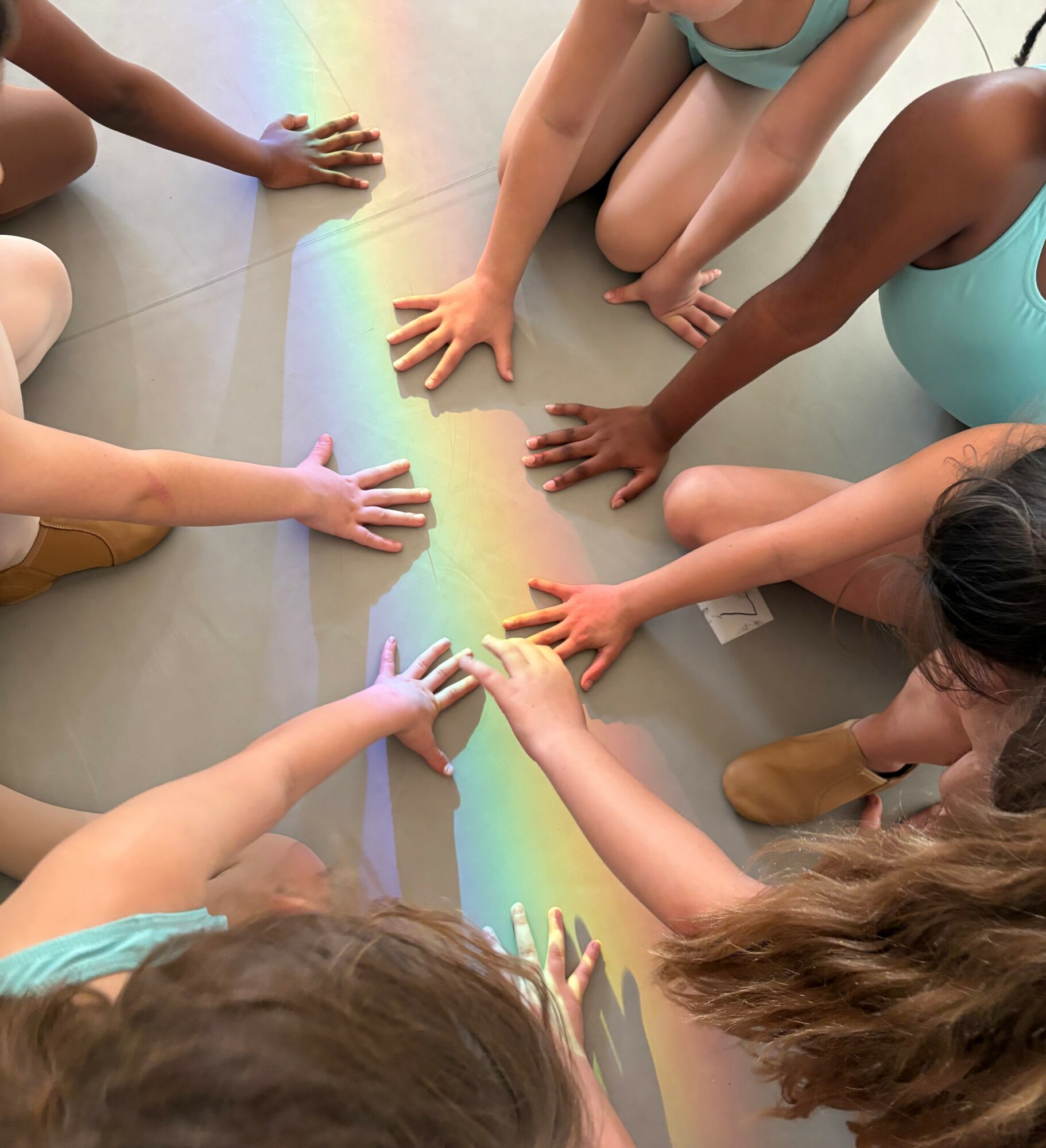 Children's hands touching a rainbow-colored light on the floor, seen from above, with some children kneeling or sitting.
