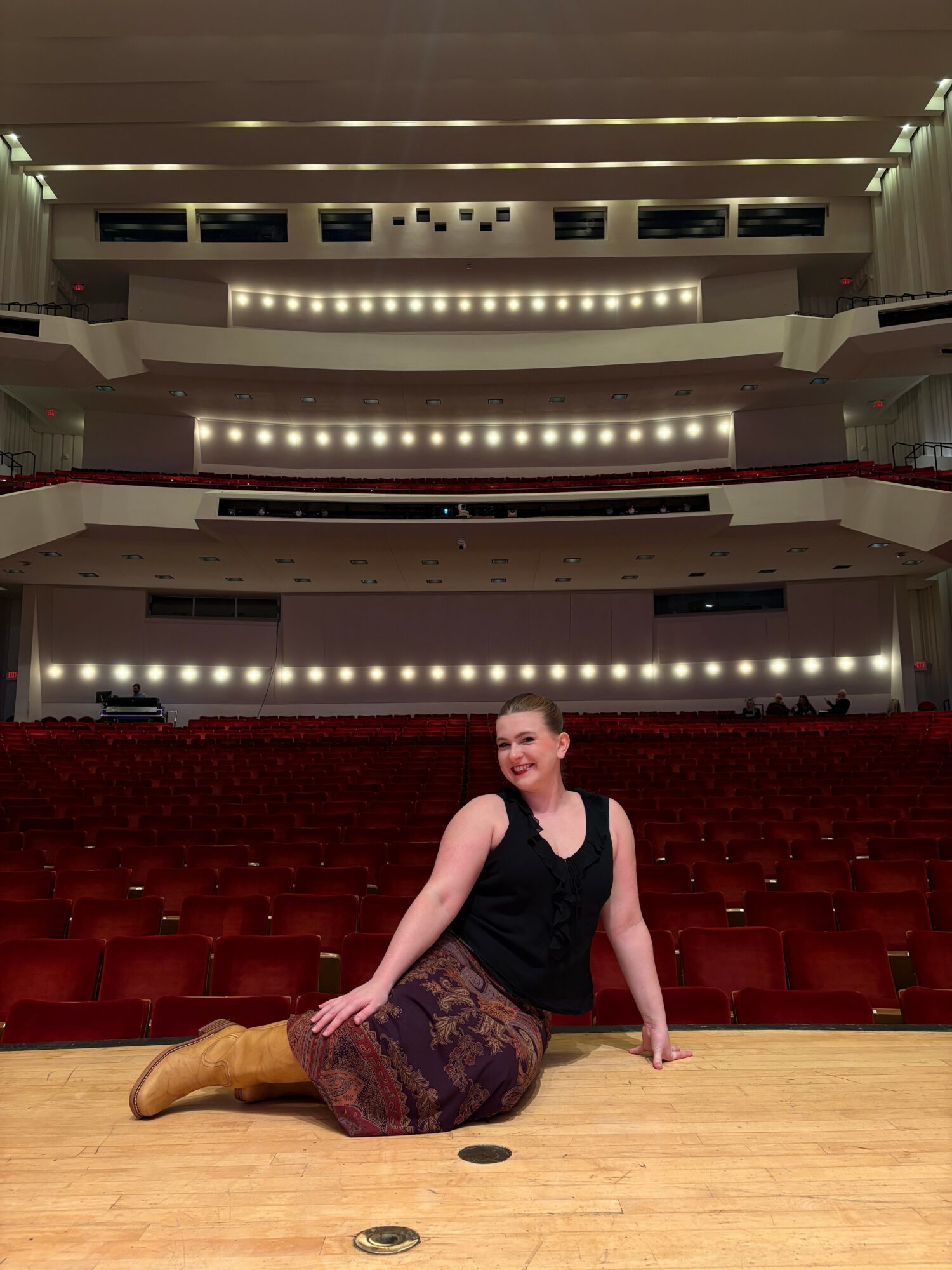 Woman sitting on stage in an empty theater, smiling, with multiple balcony levels behind her.