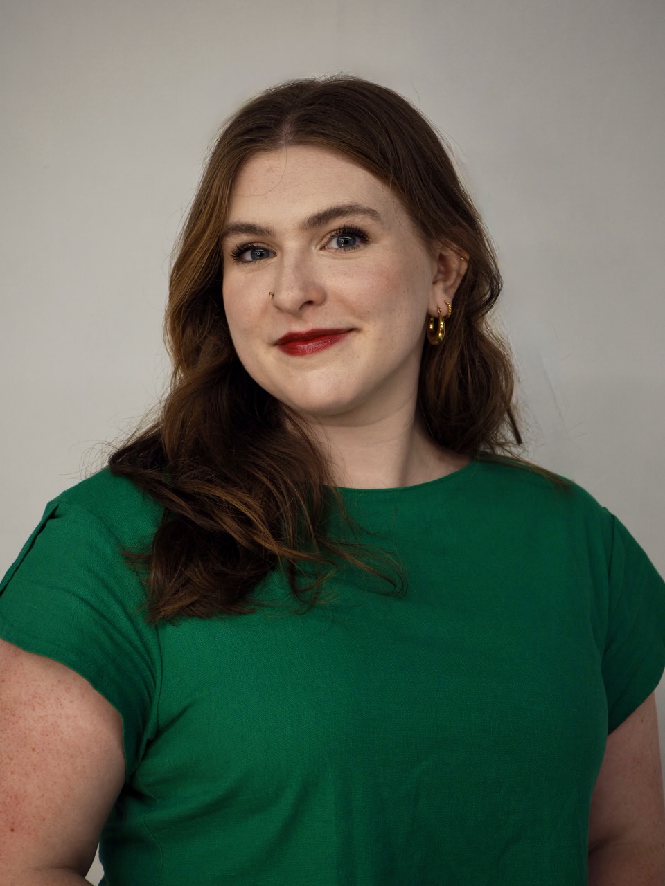 Young woman with long brown hair, red lipstick, wearing a green shirt and gold earrings, smiling at the camera.