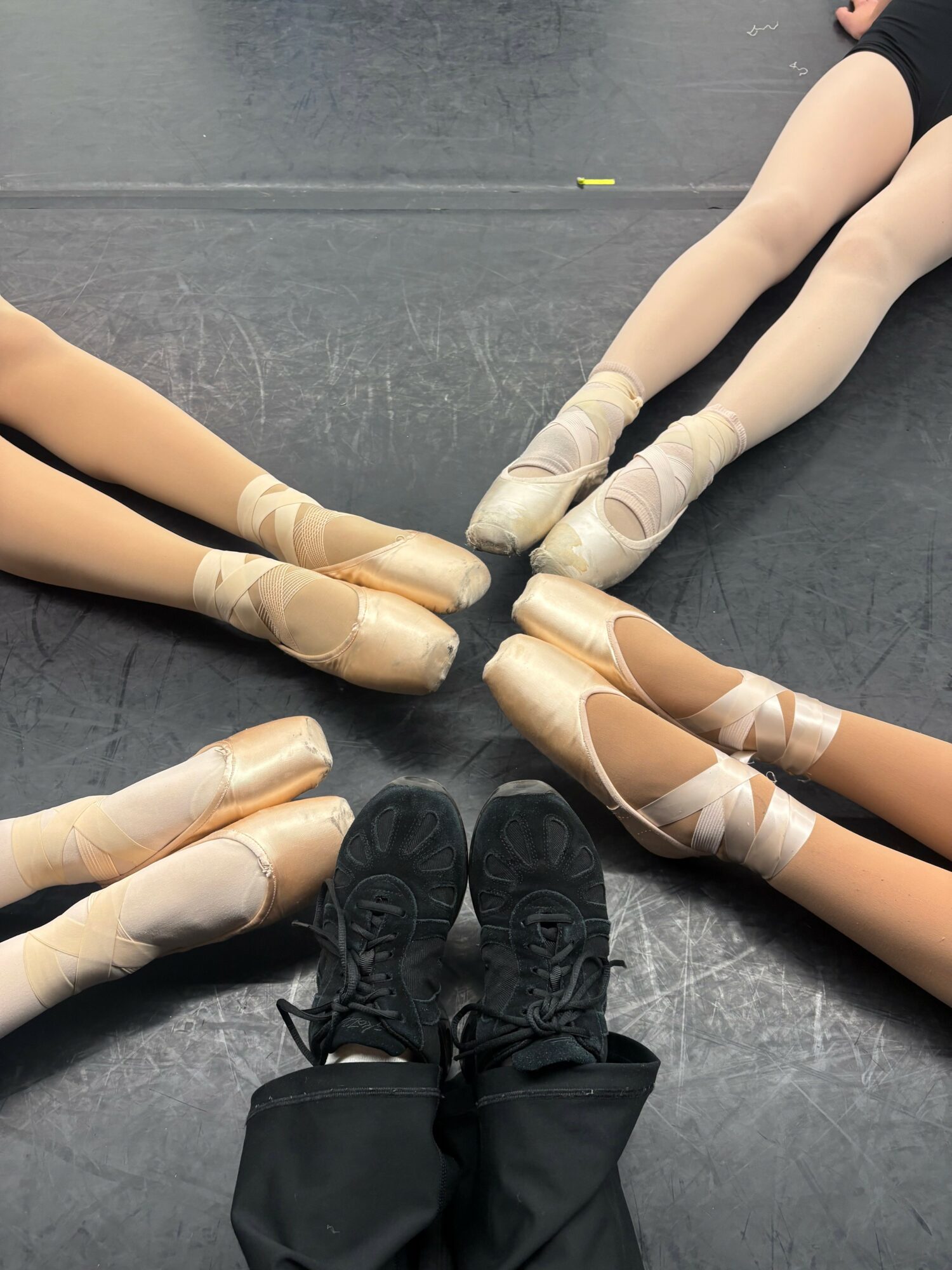 Four ballet dancers' legs and feet in pointe shoes form a circle on a dance floor, with a person's black shoes in the center.