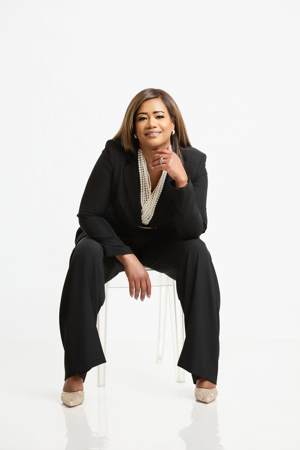 Woman sitting on a transparent chair, wearing a black suit, with a confident expression, against a white background.