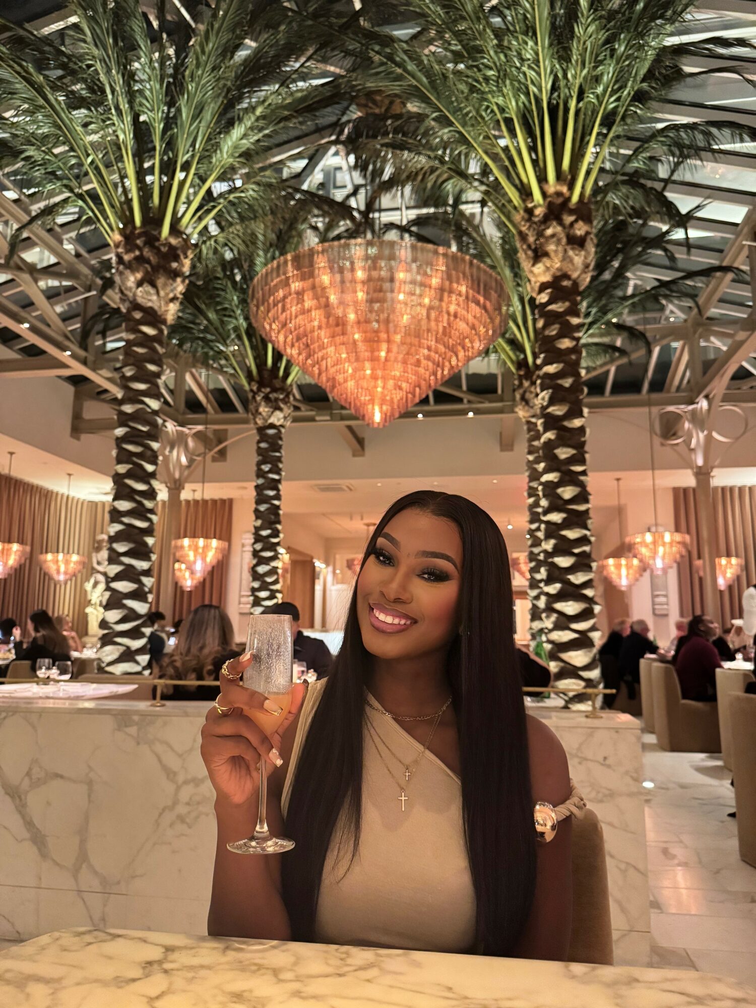 Woman smiling and holding a glass in a luxurious indoor setting with palm trees and decorative lighting.