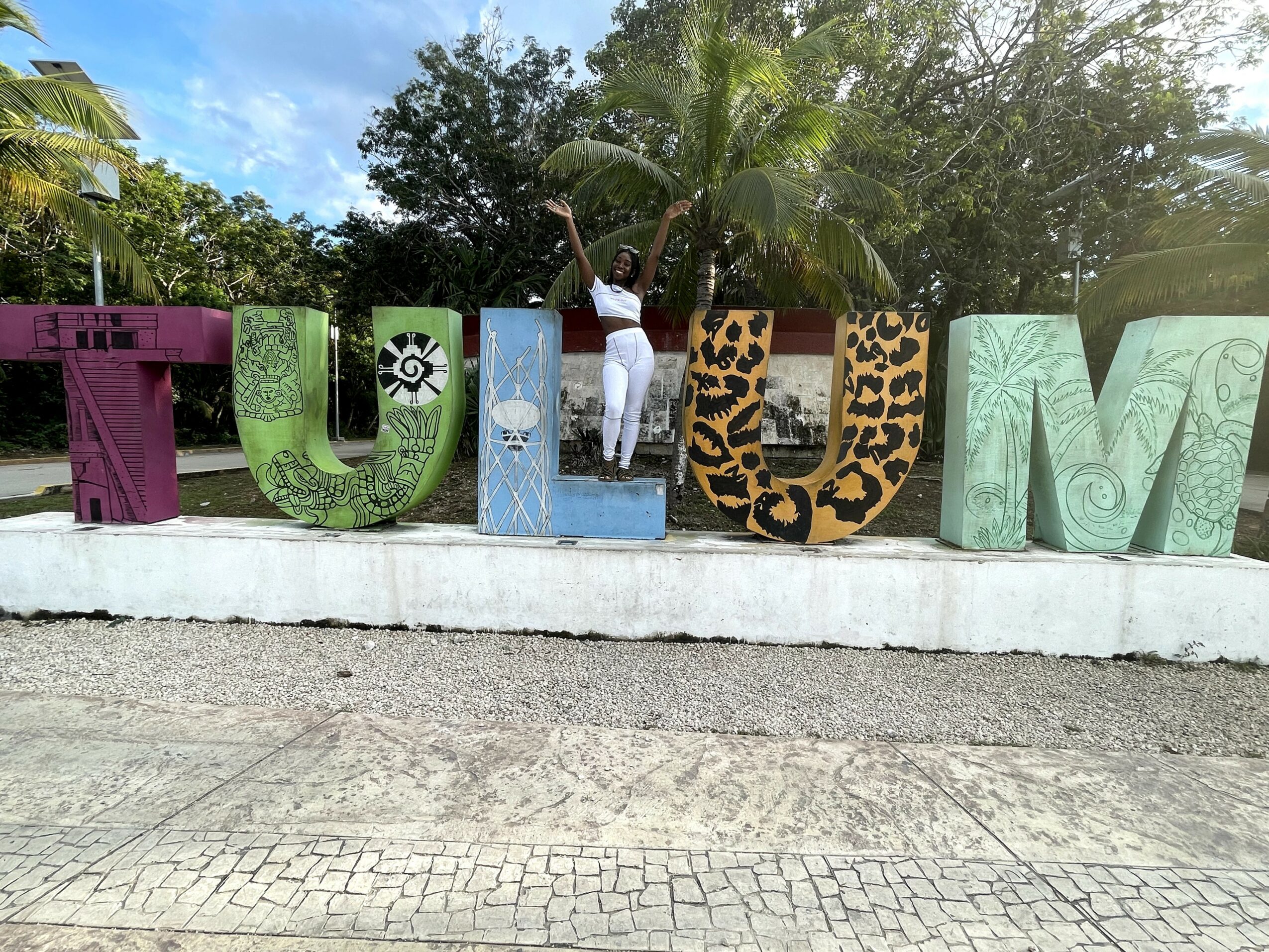 Person standing behind colorful large letters spelling 'TULUM' outdoors with trees and blue sky in background.
