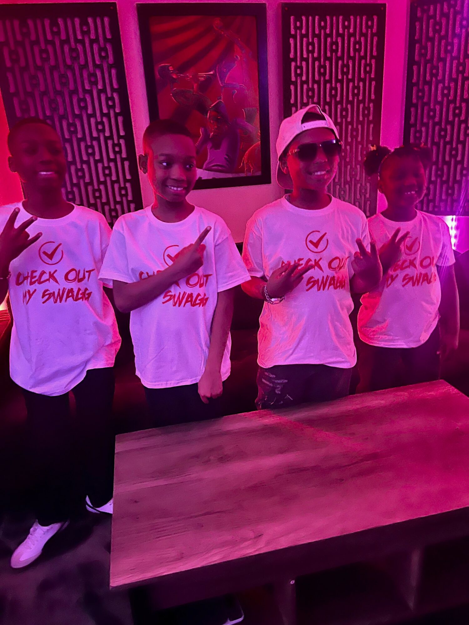Four children wearing matching white T-shirts with red text, standing indoors with pink lighting, smiling and making peace signs.
