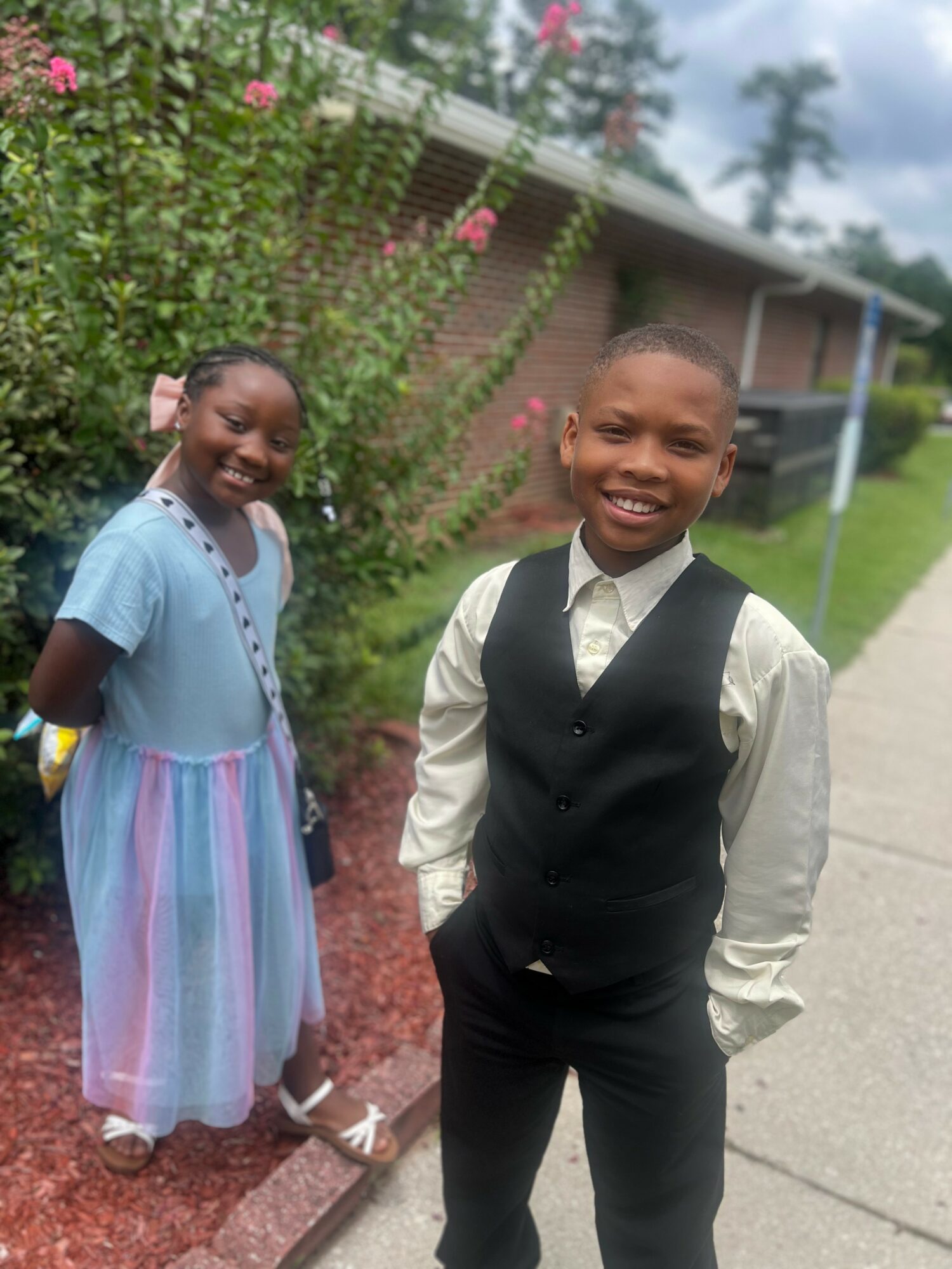 Two children, a girl in a pastel dress and a boy in a vest and tie, stand outdoors near a sidewalk with greenery and a building in the background.