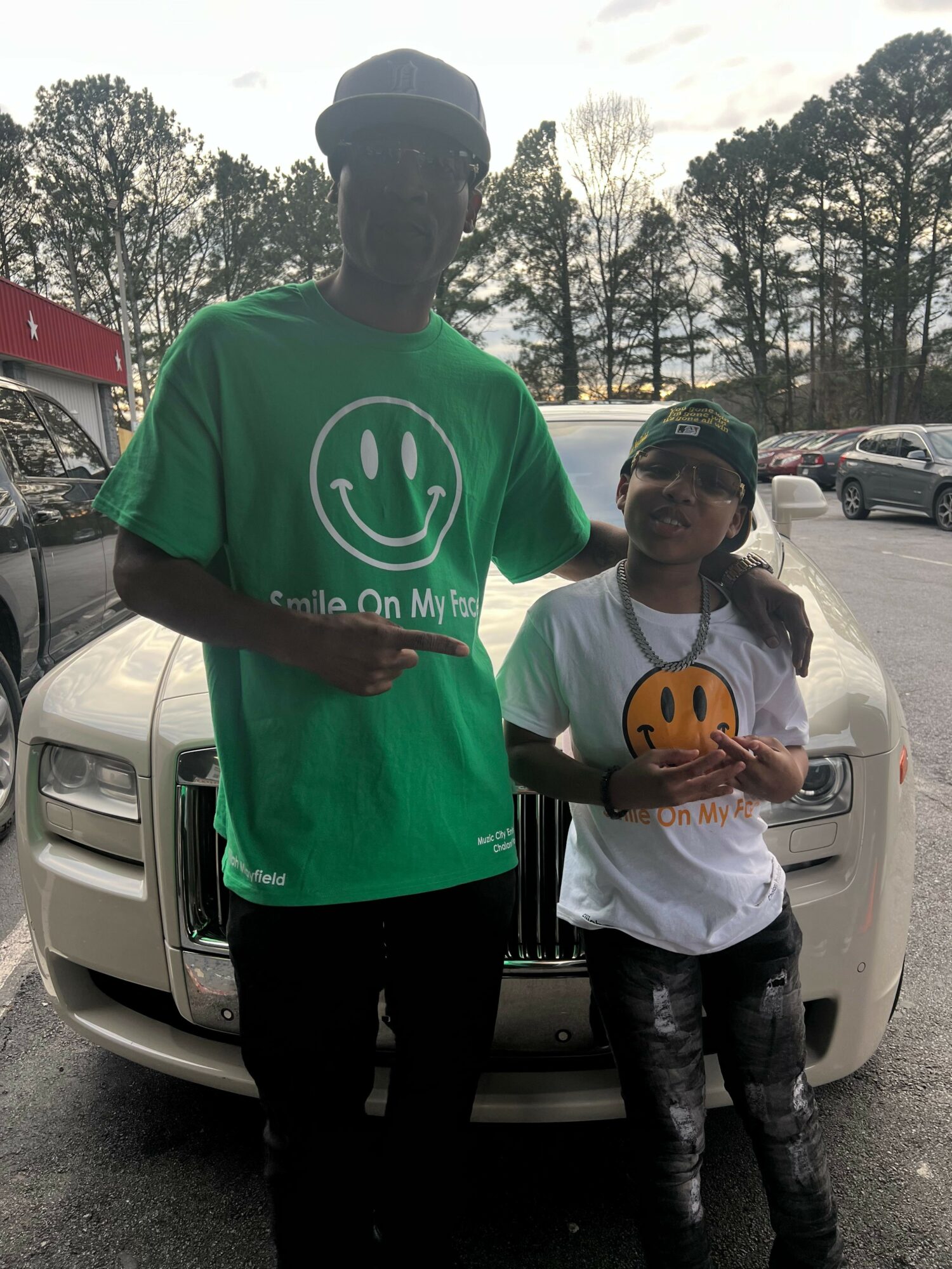 Two people standing outdoors near a white vehicle, one wearing a green shirt and the other a white shirt, holding a cake.