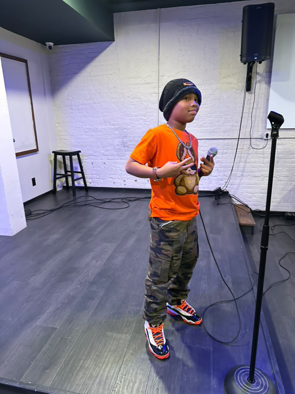 Child wearing an orange shirt, camouflage pants, and sneakers, standing on a stage with a microphone, in a room with white walls.