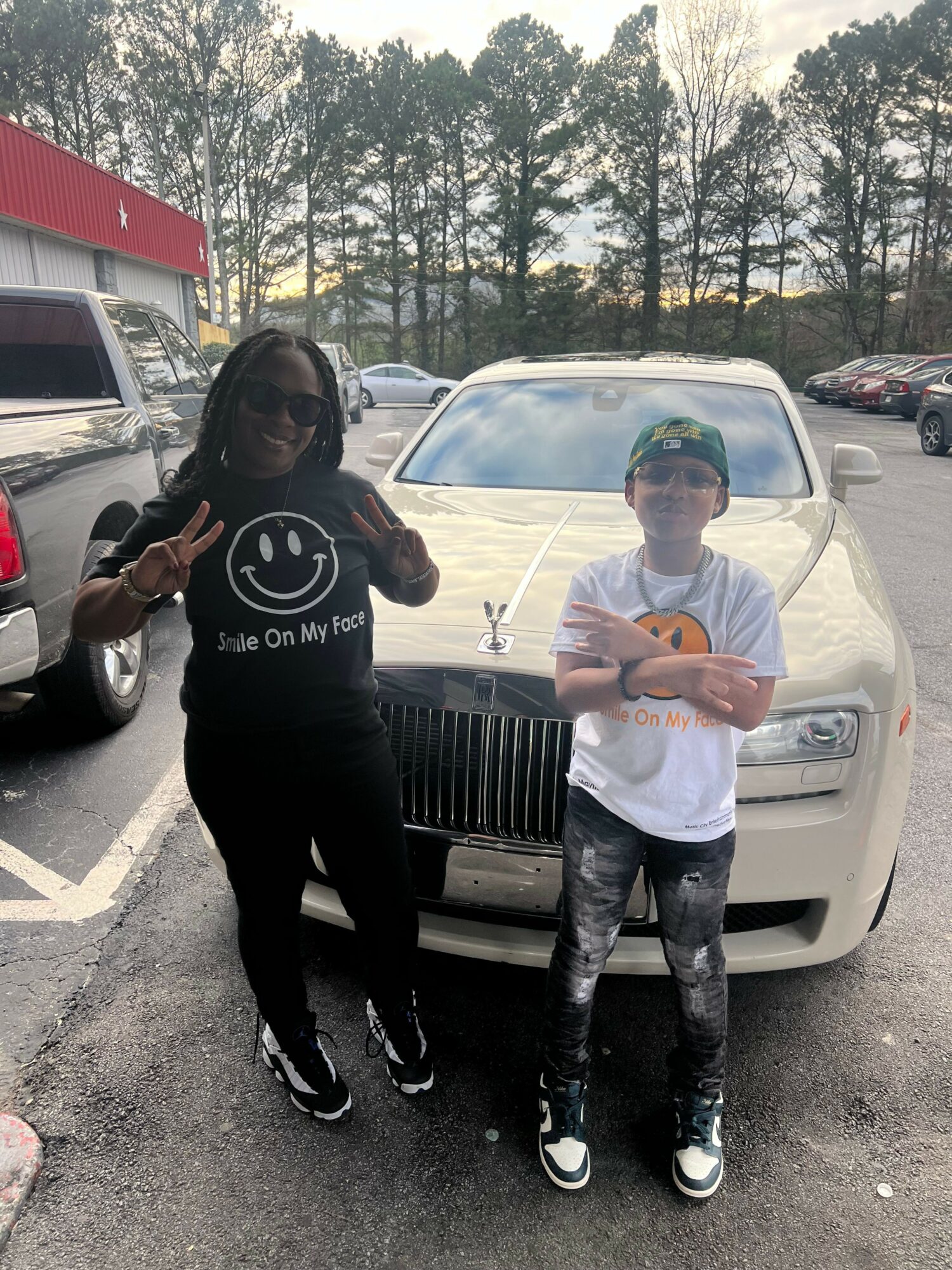 Two people standing in front of a beige car in a parking lot, one making a peace sign, trees in background.