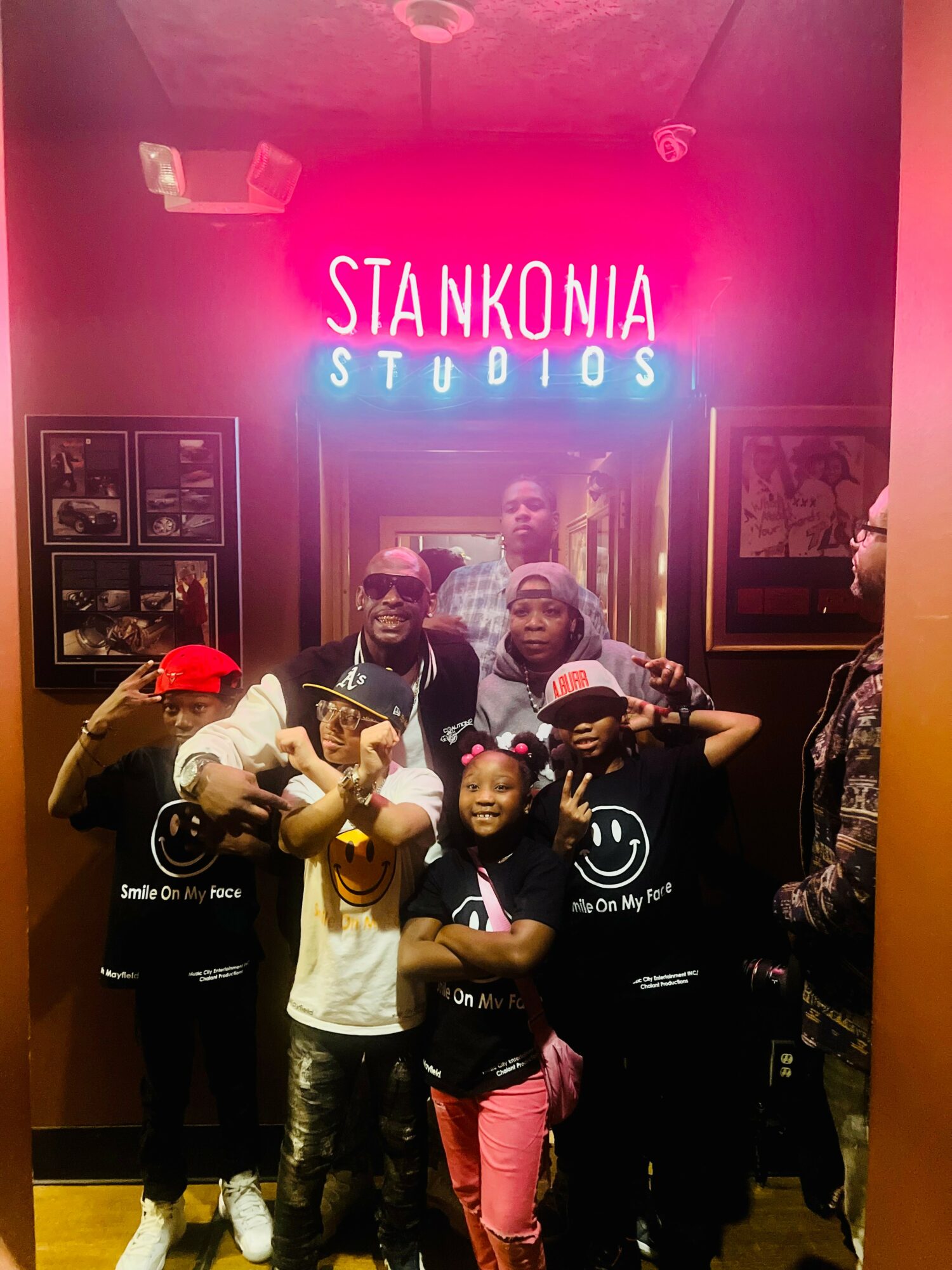 Group of children and adults posing in front of a neon sign that reads 'STANKONIA STUDIOS' inside a room with framed pictures on the wall.