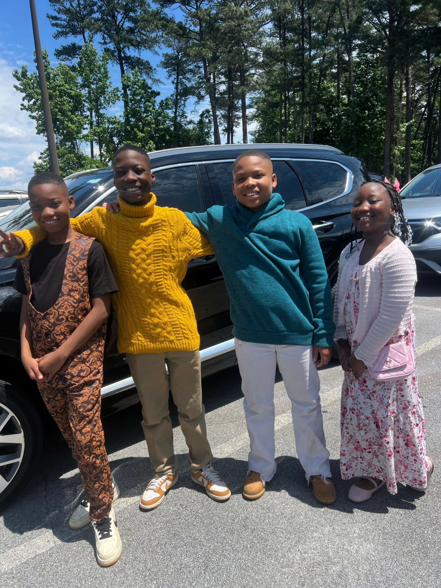 Four people standing outdoors near a car, with trees in the background.