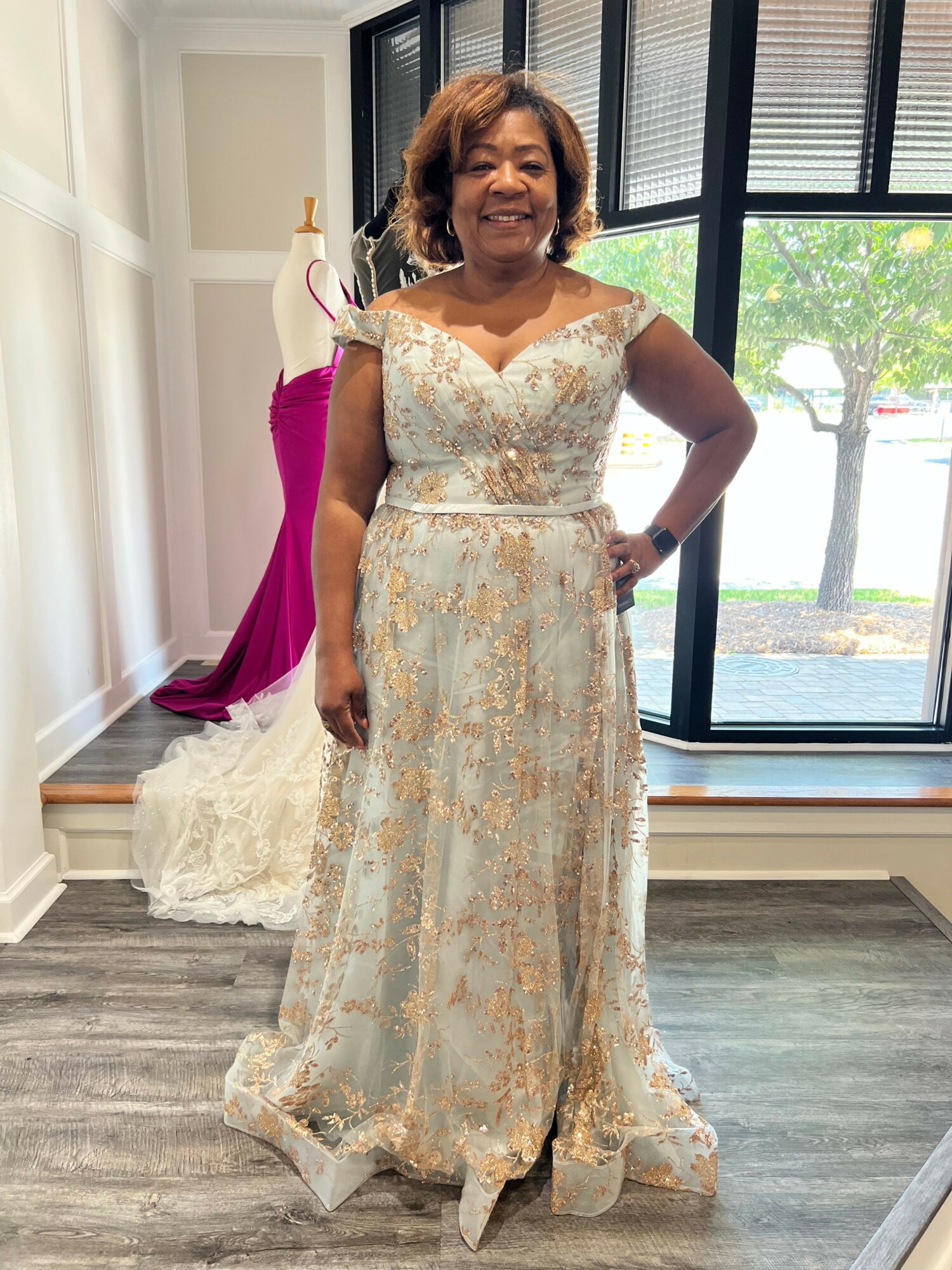 Woman in a long, light-colored lace dress standing indoors near a window with a view outside.