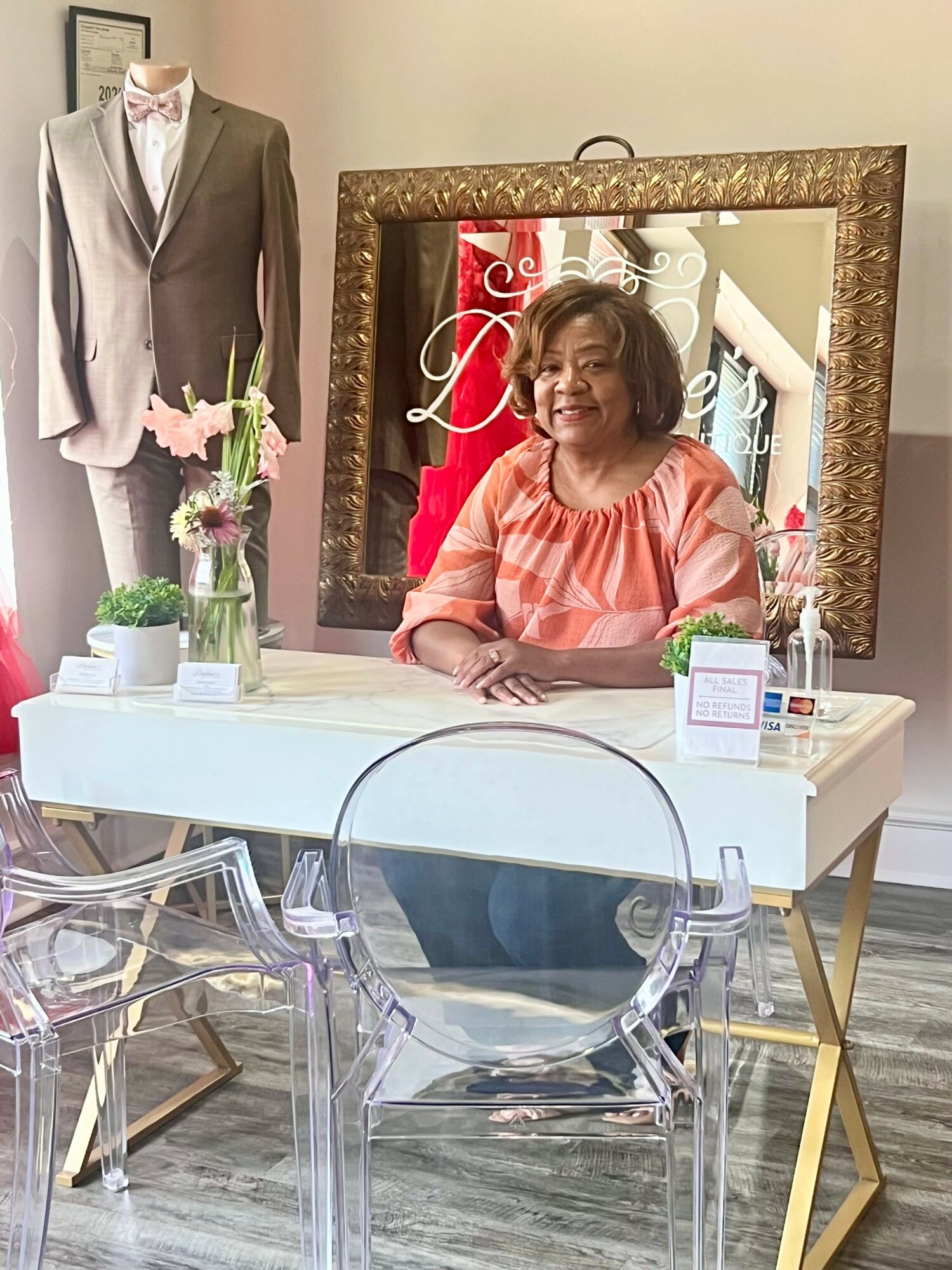 Woman sitting at a white table with a mirror behind her, a suit on a mannequin, and flowers on the table.