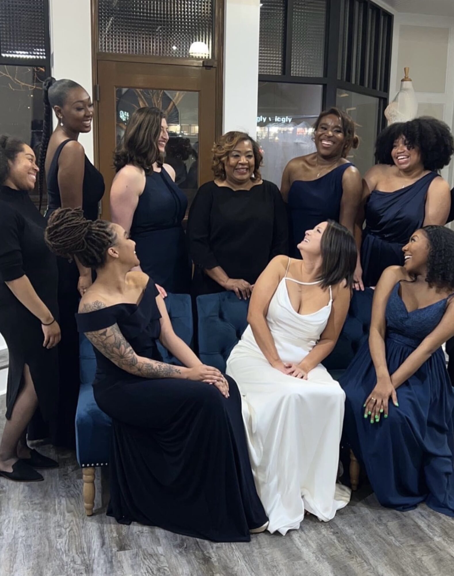 Group of women in formal dresses gathered indoors, smiling and engaging with each other, with a mannequin in the background.